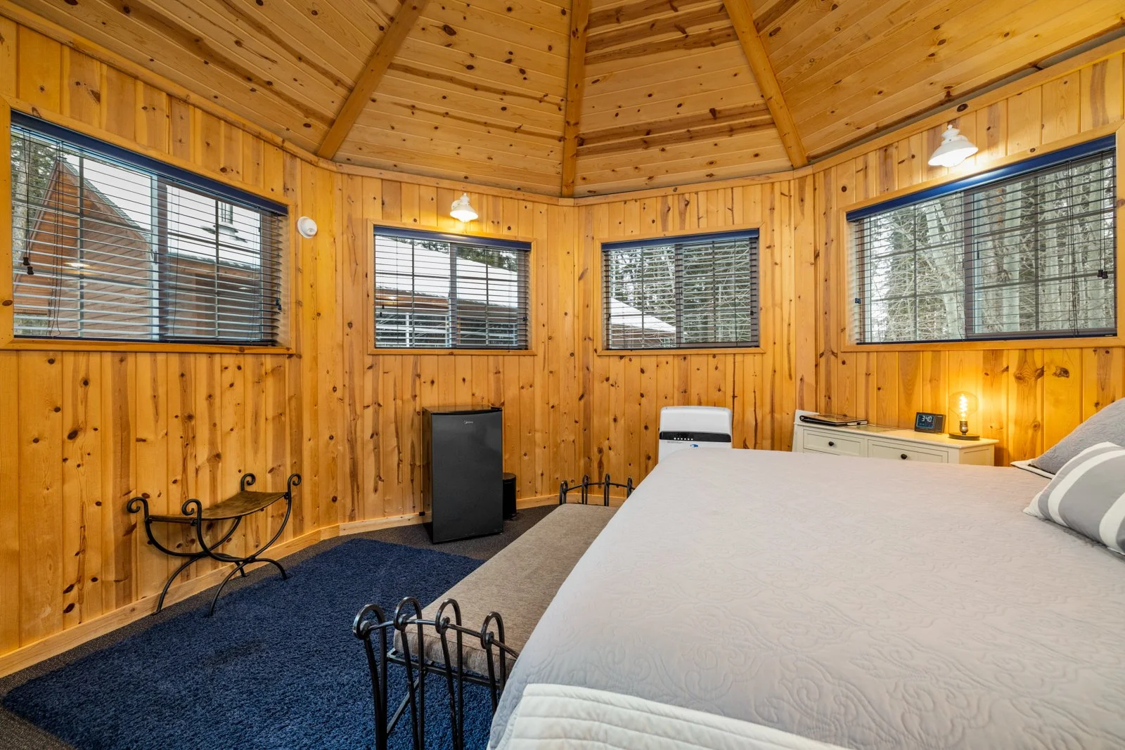 A cozy bedroom with wooden paneled walls and ceiling, large windows with blinds, a bed with pillows, a small white nightstand with a clock and a lamp, a black mini fridge, a small white heater, and a blue rug.