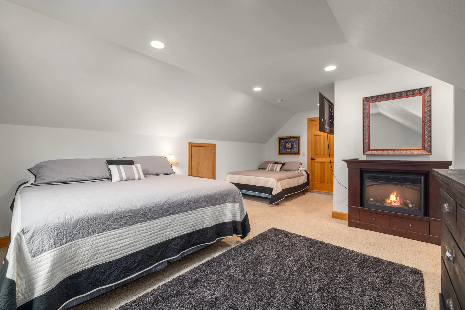 Bedroom with two beds, one large and one small, a fireplace with a mirror above, a wall-mounted TV, and a small door on the wall, with beige carpet and recessed lighting.