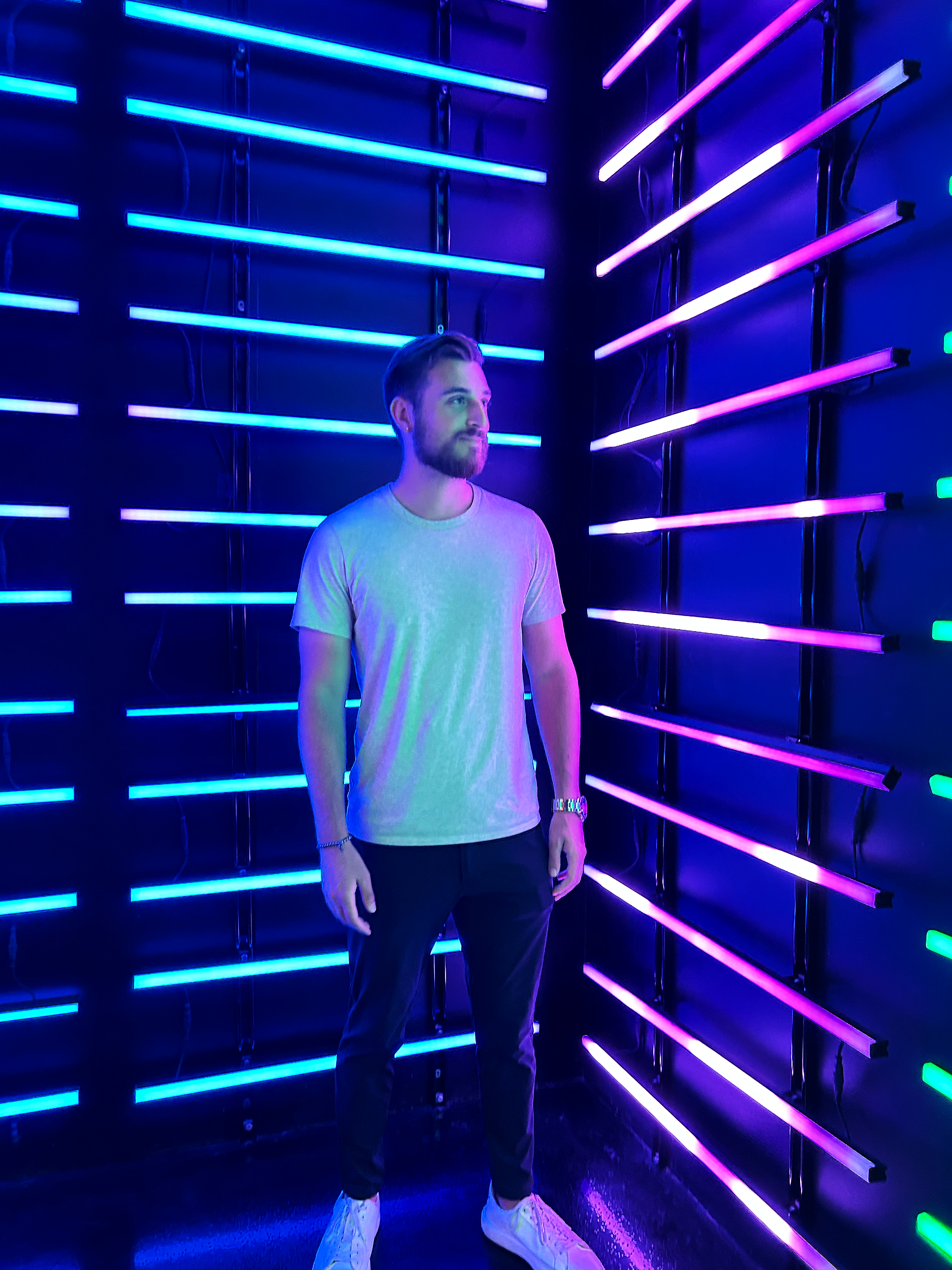 A man with a beard in casual clothes stands in front of neon blue and pink lights on a dark wall. The lights are arranged in horizontal lines on both sides of the corner. The man is looking slightly to his right with a neutral expression.