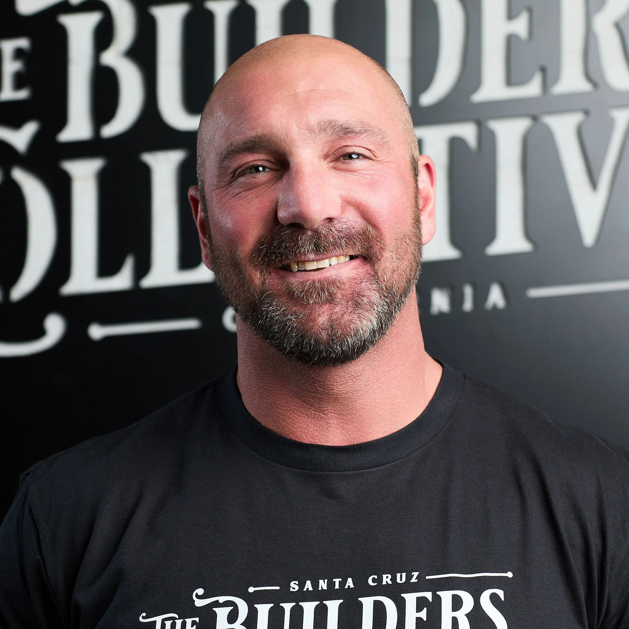 A man with a beard and shaved head smiling in front of a black background with white text that says 'The Builders Santa Cruz'.
