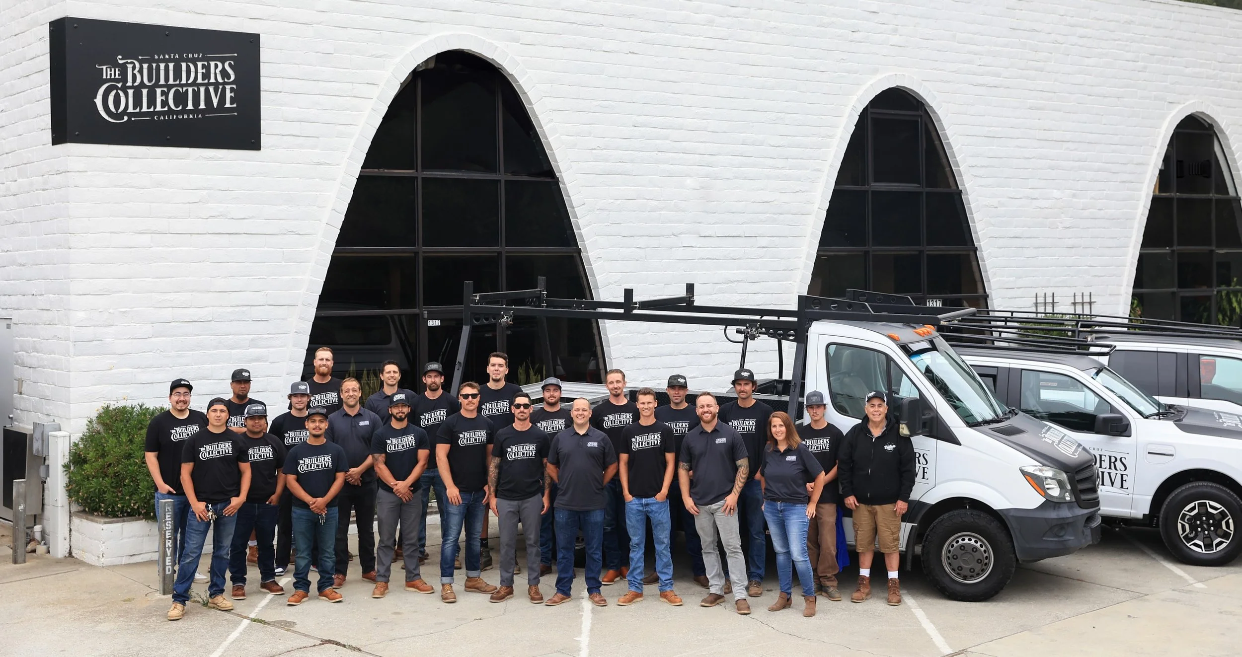 Group photo of workers standing in front of a white brick building with arched windows, some wearing black t-shirts and others in navy uniforms, with company vehicles parked nearby.