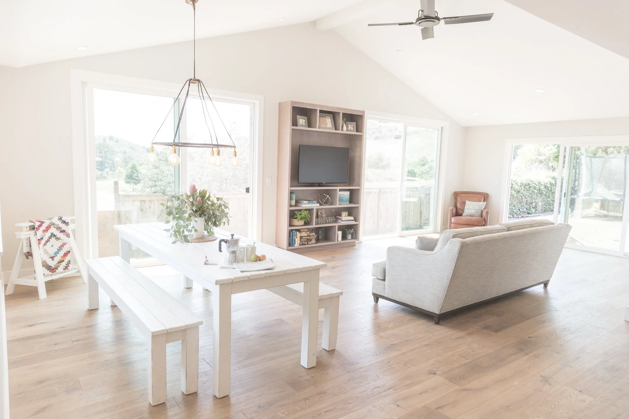 Bright living room with a white sofa facing a wooden entertainment center holding a flat-screen TV, large windows and sliding glass doors letting in natural light, a dining table with a bench, and a ceiling fan.