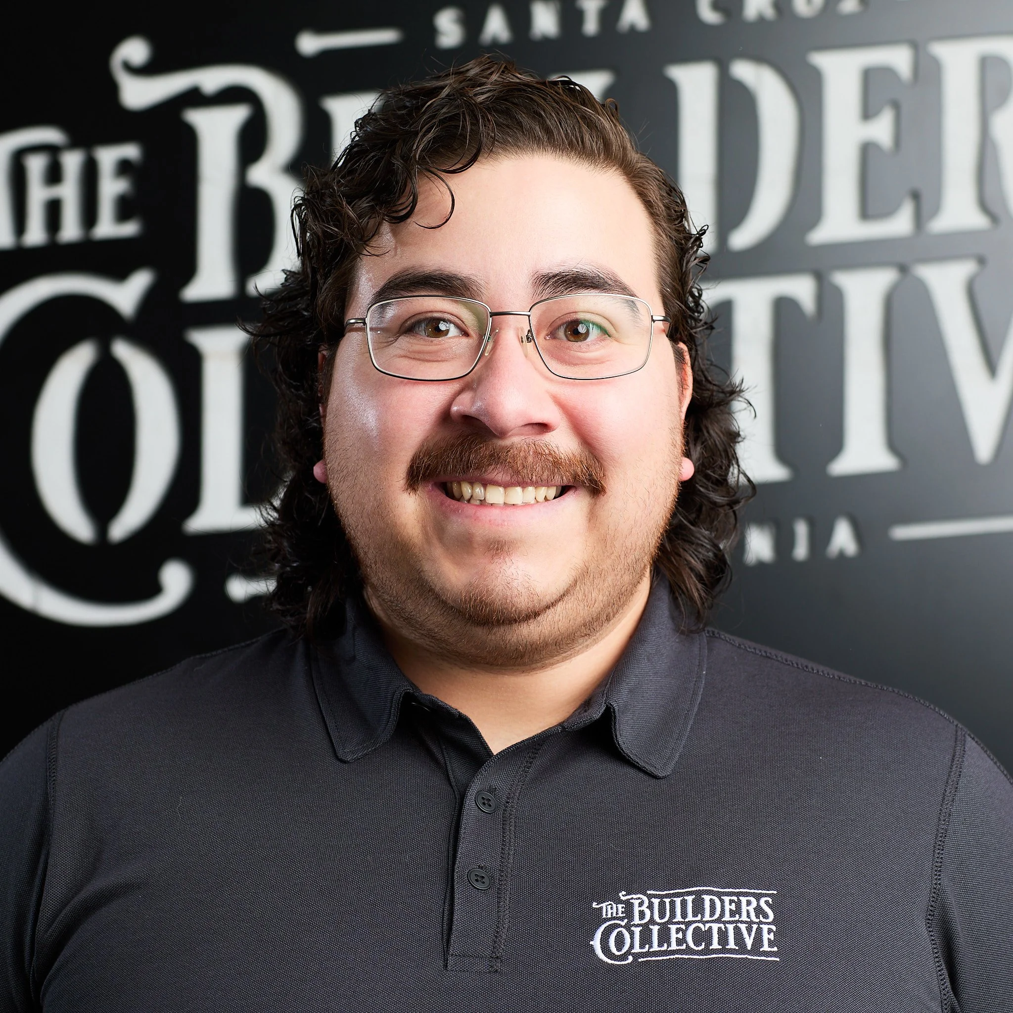 A man with glasses and a beard smiling in front of a black wall with white text that reads 'The Builders Collective'.