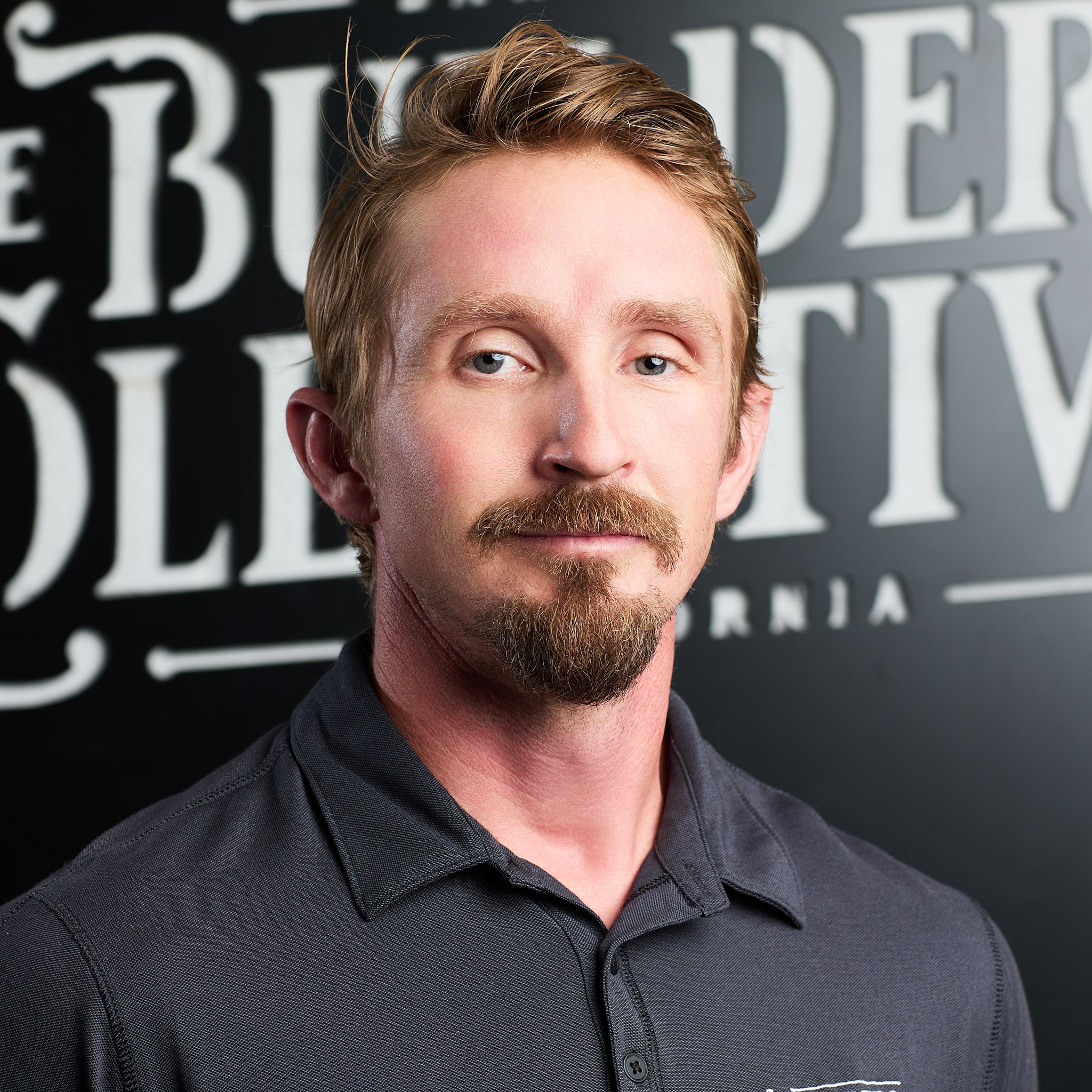 Portrait of a man with ginger hair and a beard, wearing a black collared shirt, standing in front of a black background with white large text.