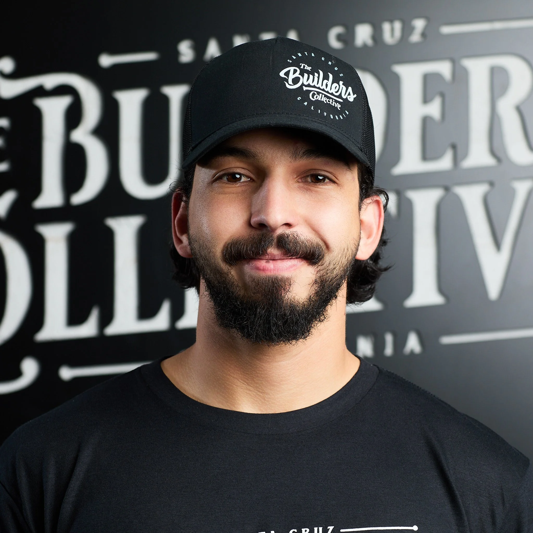 A man with a mustache and beard wearing a black cap and black T-shirt, standing in front of a black and white sign that reads 'The Builders Collective California'.