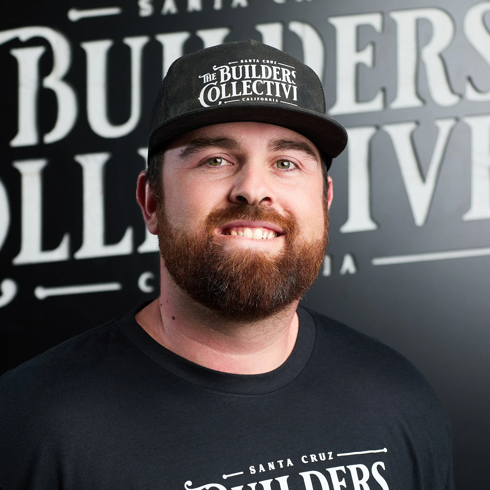 Man with a beard wearing a black cap and black T-shirt, standing in front of a black and white sign that reads 'The Builders Collective'.