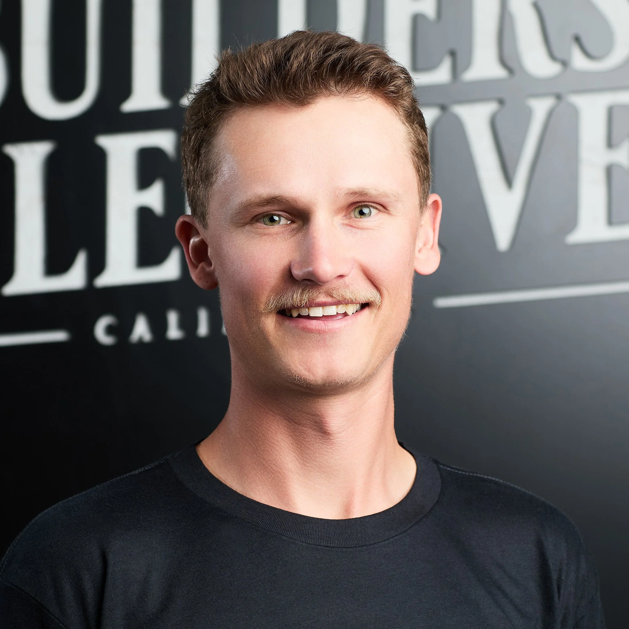 A young man with short, light brown hair, a mustache, and a black t-shirt, smiling in front of a black background with white lettering.