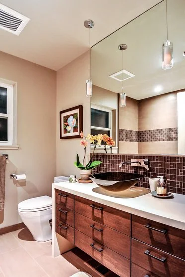 A modern bathroom with beige walls, a large mirror, and a wooden vanity with a white countertop. Decorative items include a potted orchid, candles, and artwork of a flower on the wall. The bathroom features pendant lighting, a window, and a section of mosaic tile on the wall.