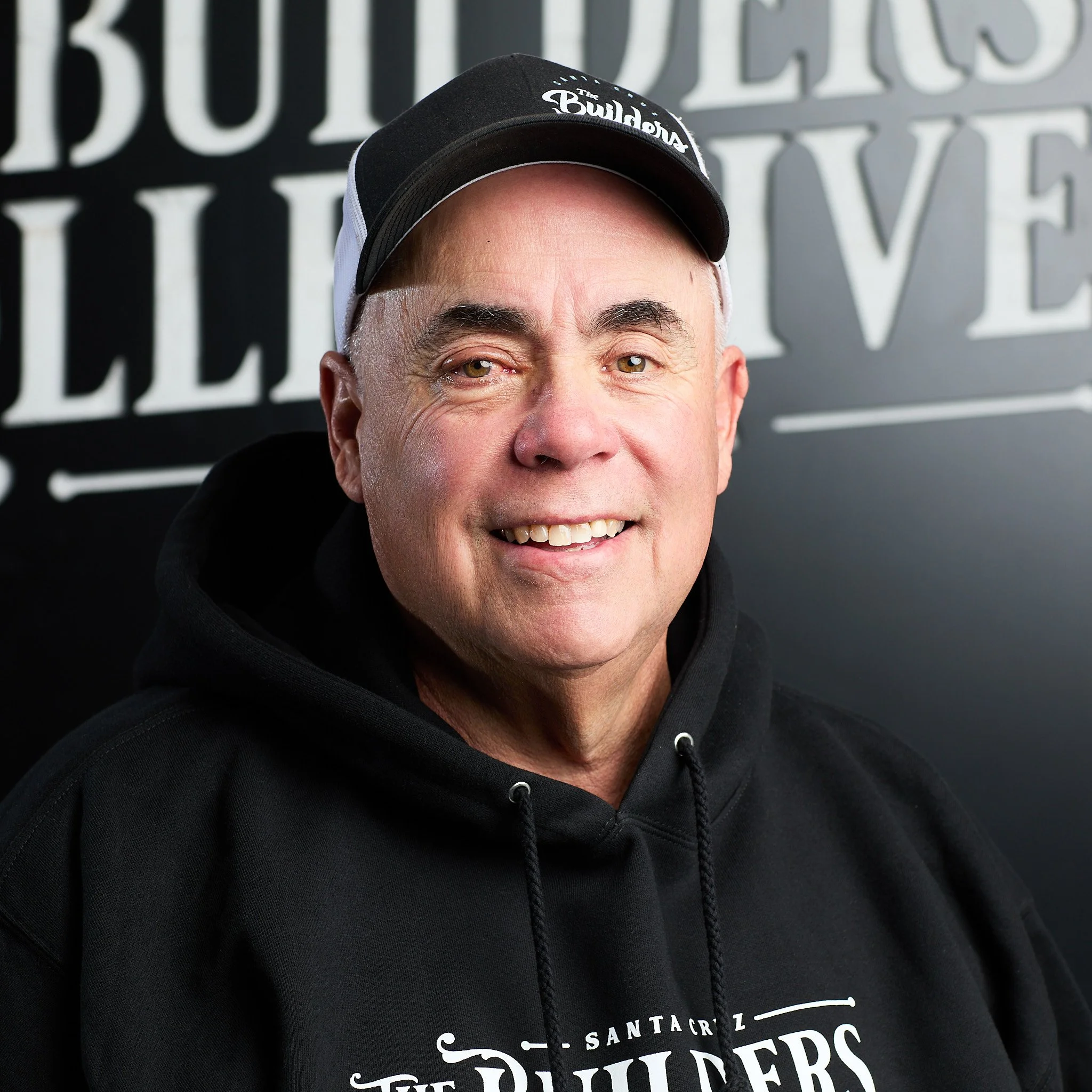 A smiling middle-aged man with gray hair wearing a black hoodie and a black and white cap, standing in front of a black background with white text.