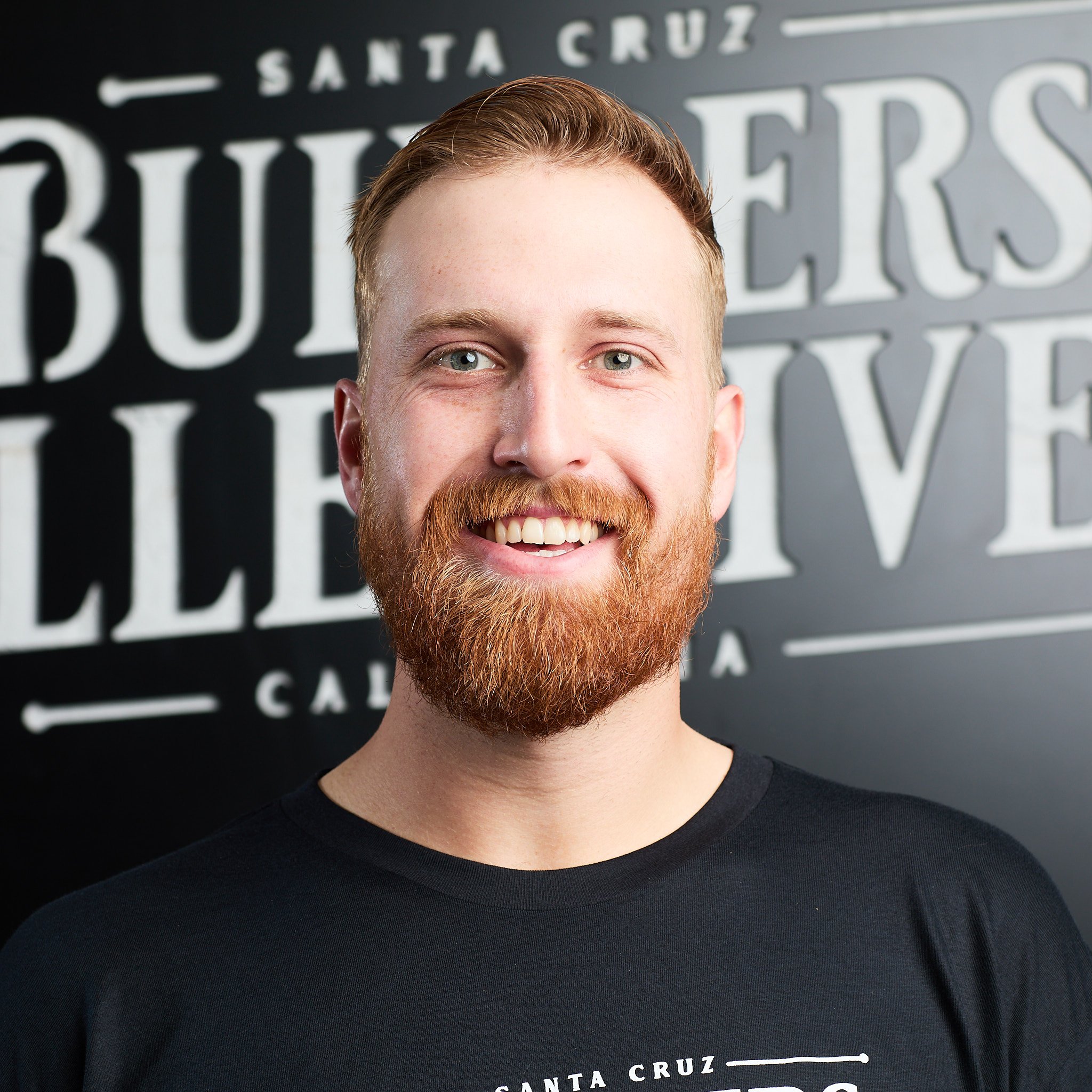 A smiling man with red hair and beard standing in front of a wall with large white text that reads 'Santa Cruz' and 'Bubbers'.