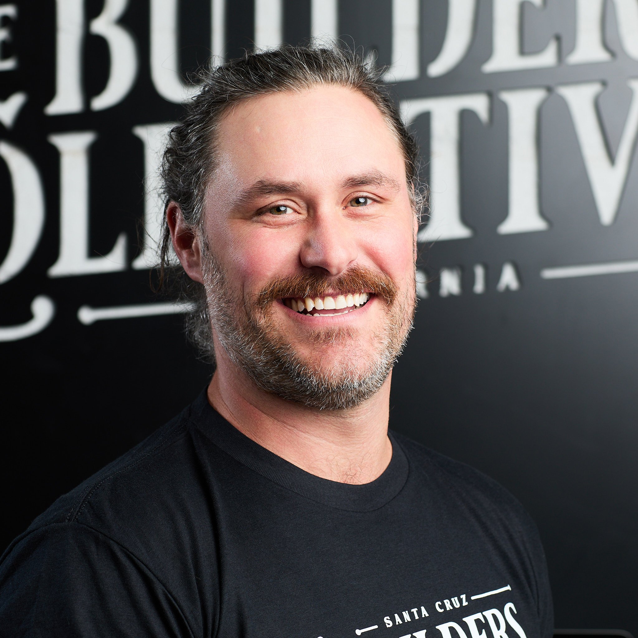 Smiling man with beard and long hair, wearing a black T-shirt, standing in front of a black wall with white text.