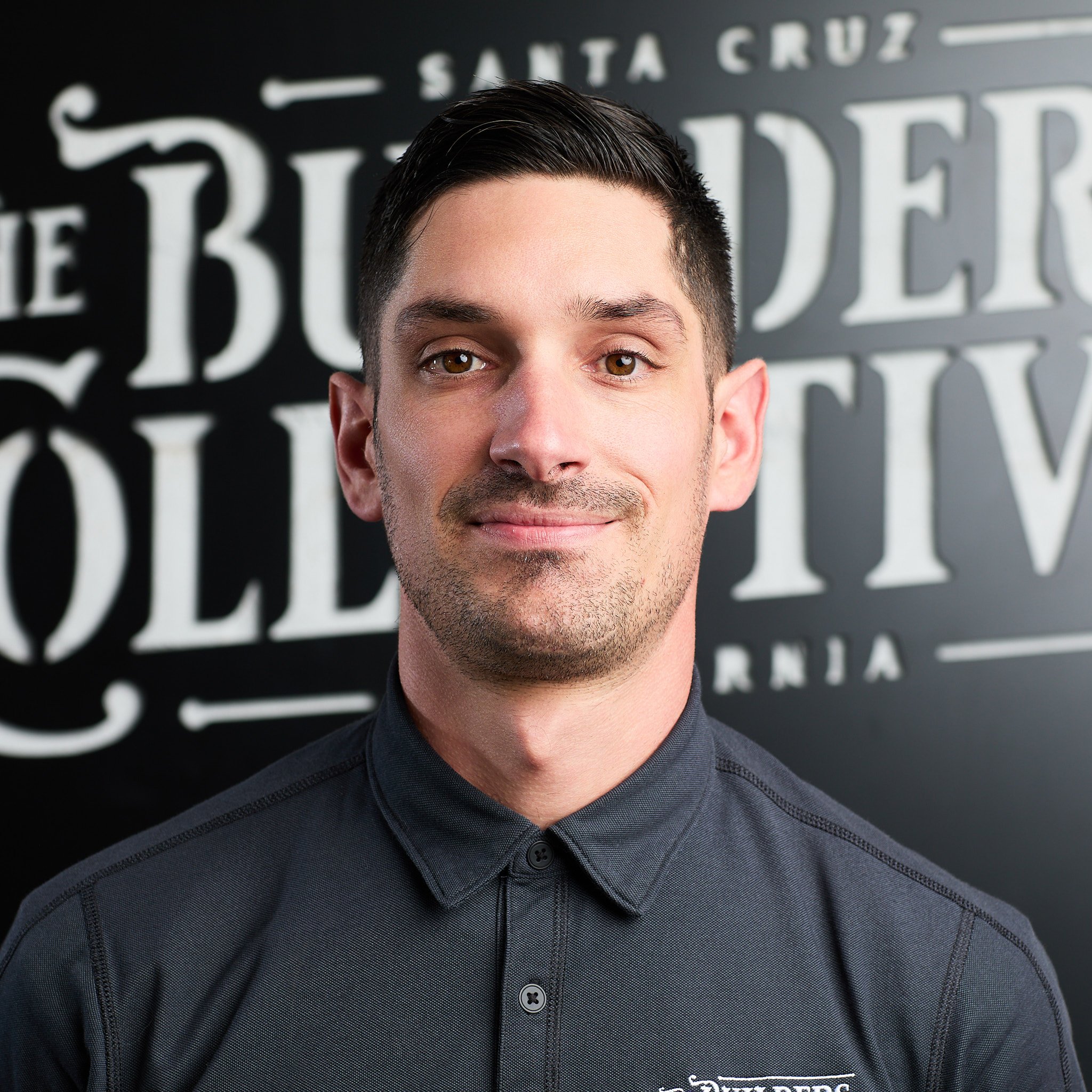 A young man with short dark hair and light skin, wearing a dark grey collared shirt, standing in front of a black background with white text.