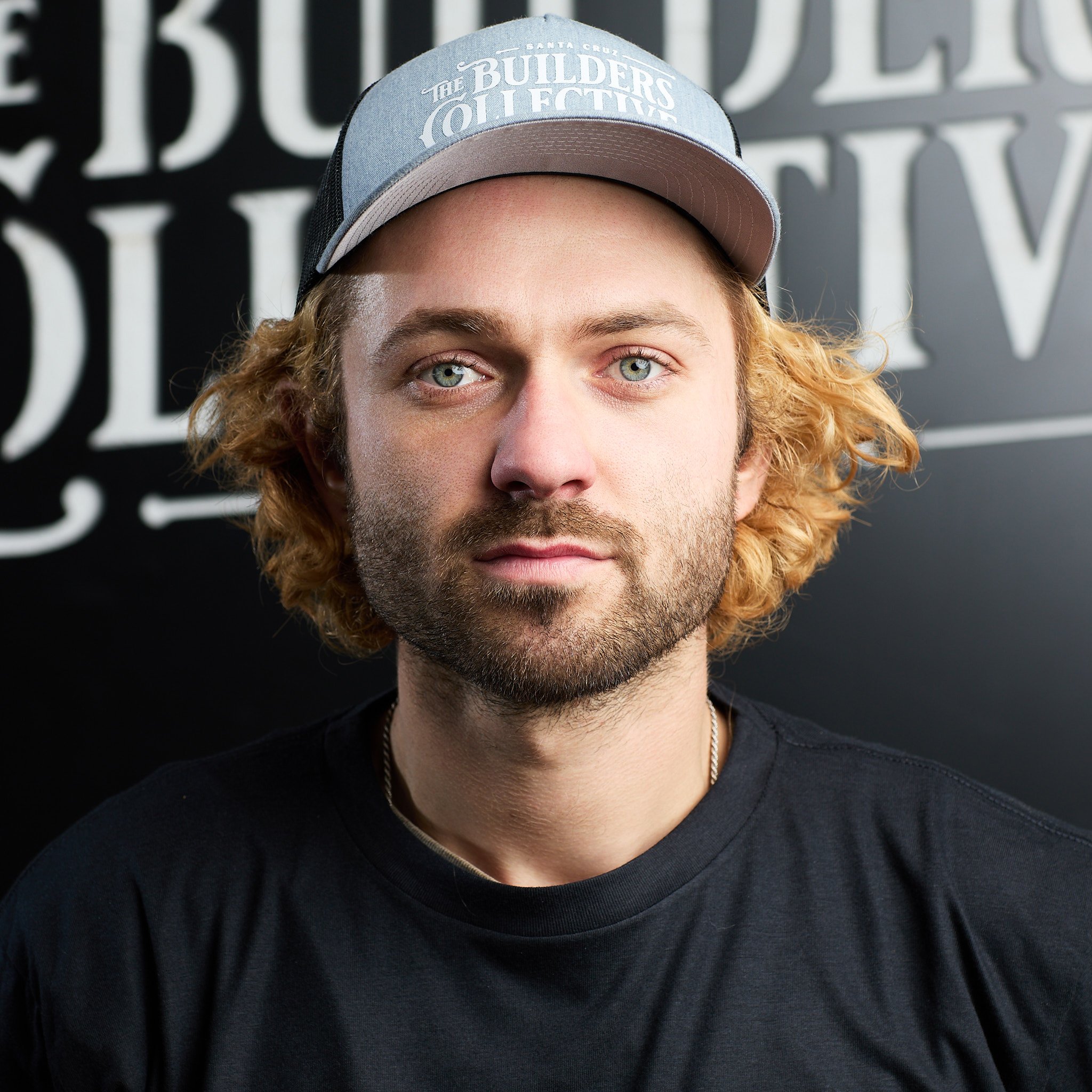 A young man with blond, curly hair and a beard wearing a gray and black cap that says "The Builders Collective" and a black T-shirt. He is in front of a black background with white lettering.
