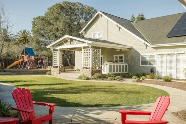 Backyard of a house with a lawn, two red Adirondack chairs, a playground set with a slide, and a house with solar panels on the roof.
