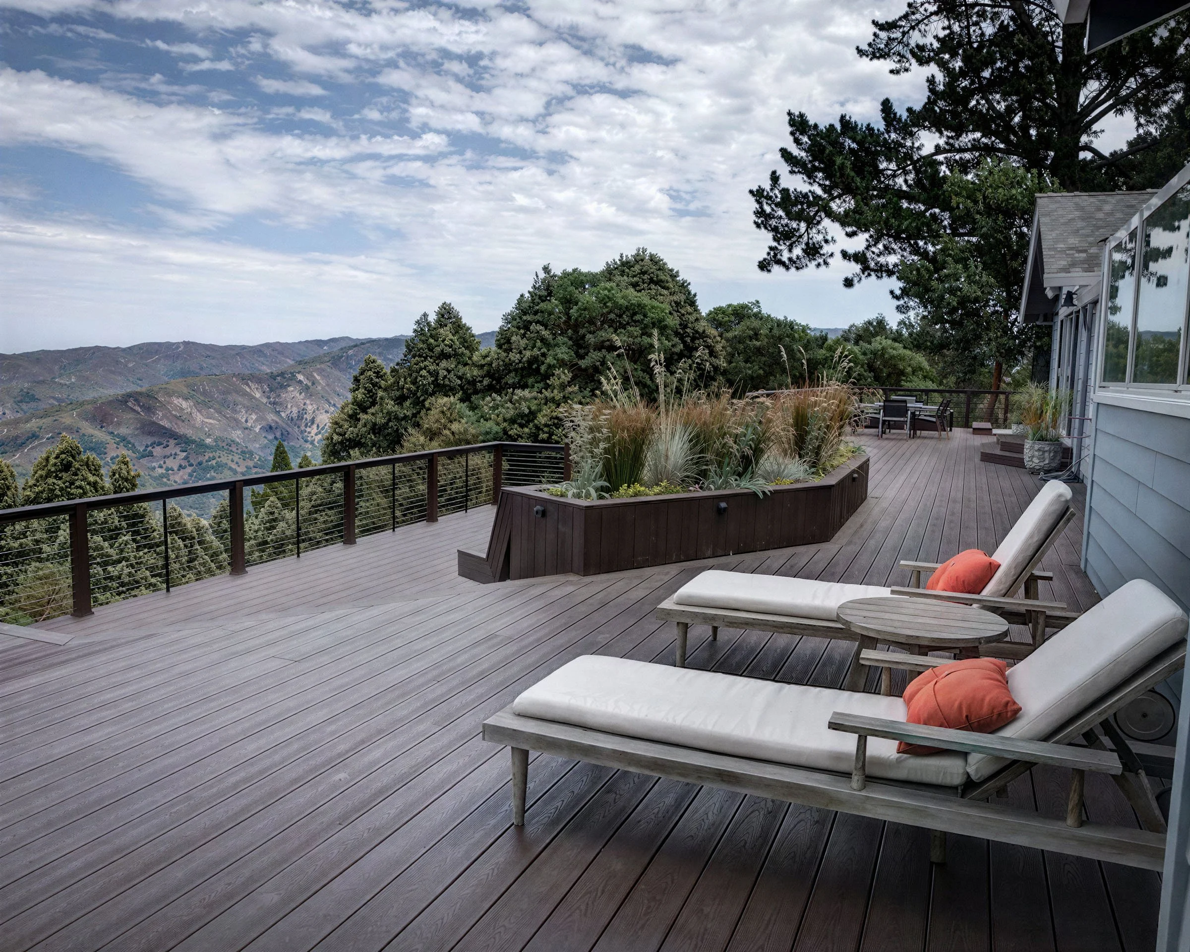 A spacious wooden deck with lounge chairs and potted plants, overlooking a mountainous landscape under a partly cloudy sky.