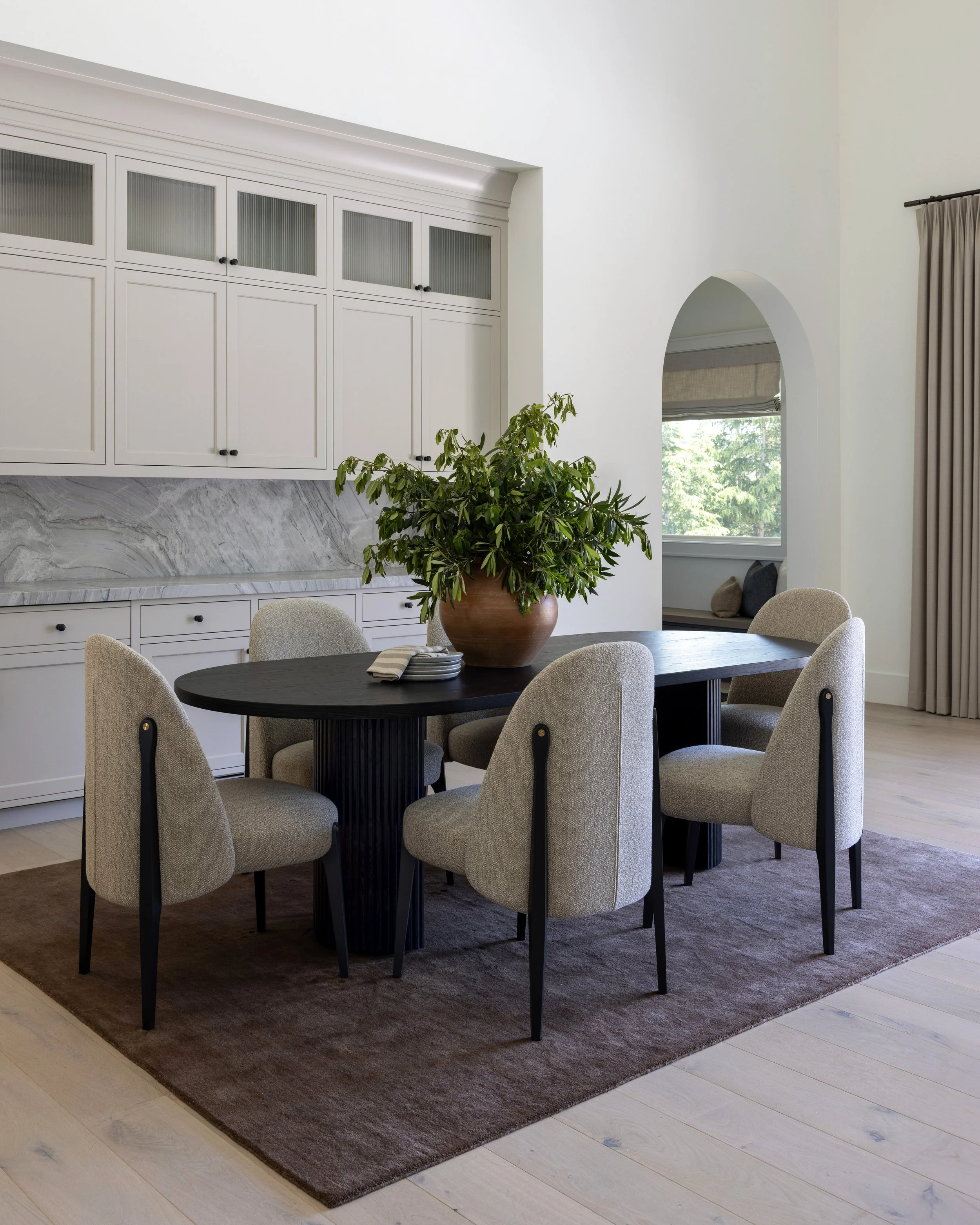 Modern dining room with a dark wood table, six beige upholstered chairs, a large potted plant, white cabinets, and an arched doorway with a view outside.