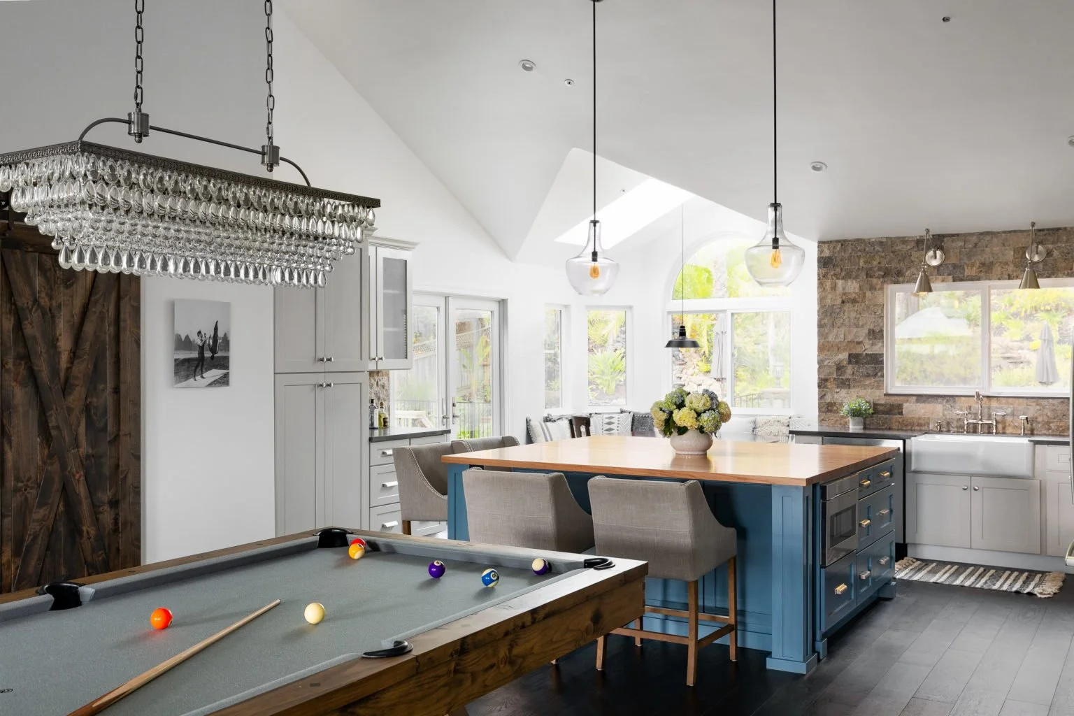 Interior of a bright, modern kitchen with a pool table in the foreground. The kitchen features a large island with a wooden top, blue base cabinets, and gray stools. There are white cabinets along the wall, a farmhouse sink, and large windows allowing natural light. A crystal chandelier hangs above the pool table, and three hanging pendant lights illuminate the island. A bouquet of flowers is on the island, and the kitchen has a mix of wood and brick accents.