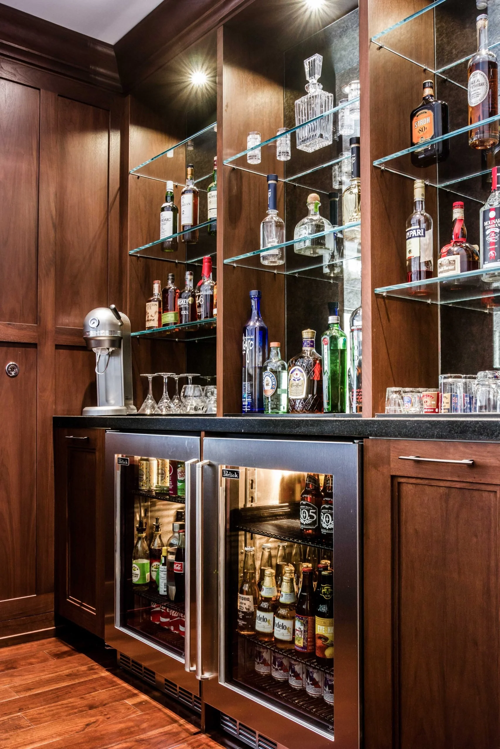 A home bar with wooden cabinetry, glass shelves holding various liquor bottles, an ice maker, and a small fridge stocked with beverages.