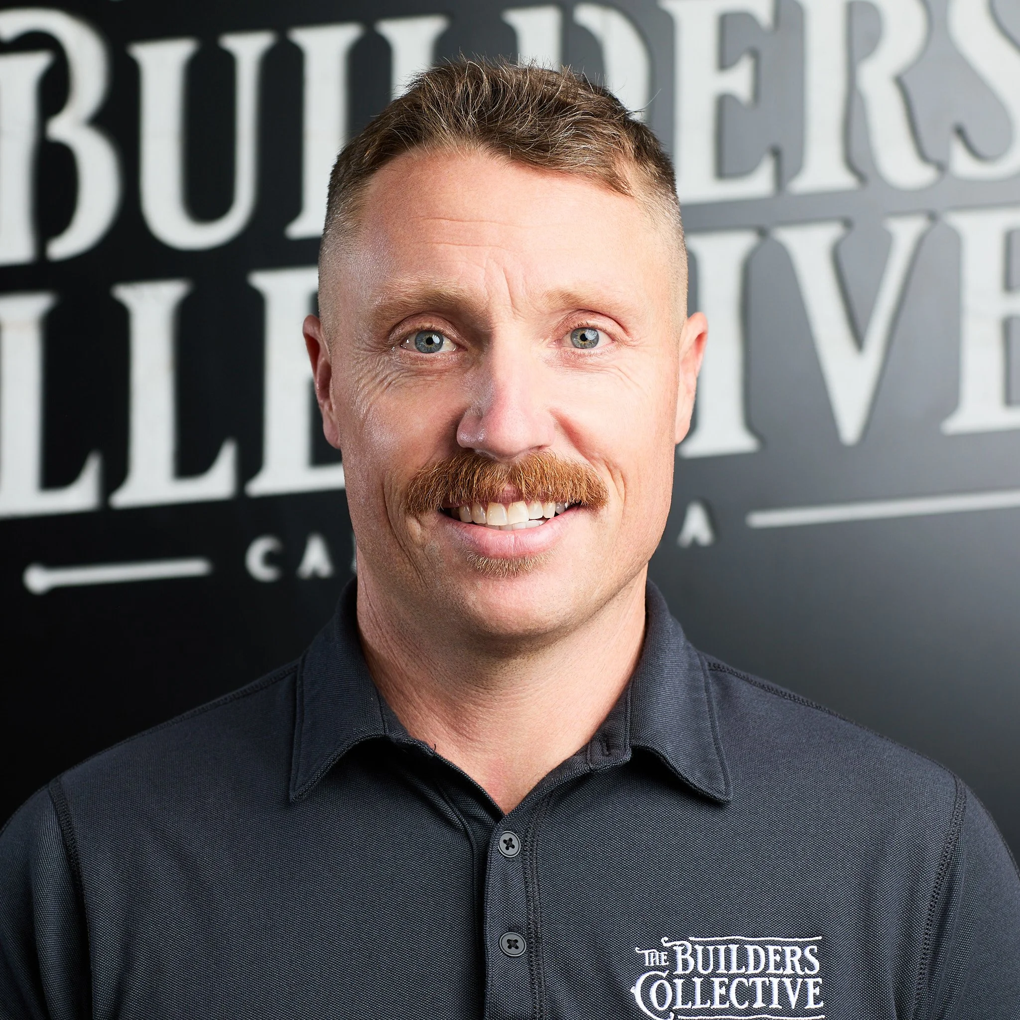 A man with short hair and a mustache smiling, wearing a black collared shirt with a logo reading 'The Builders Collective' in front of a black background with large white letters.