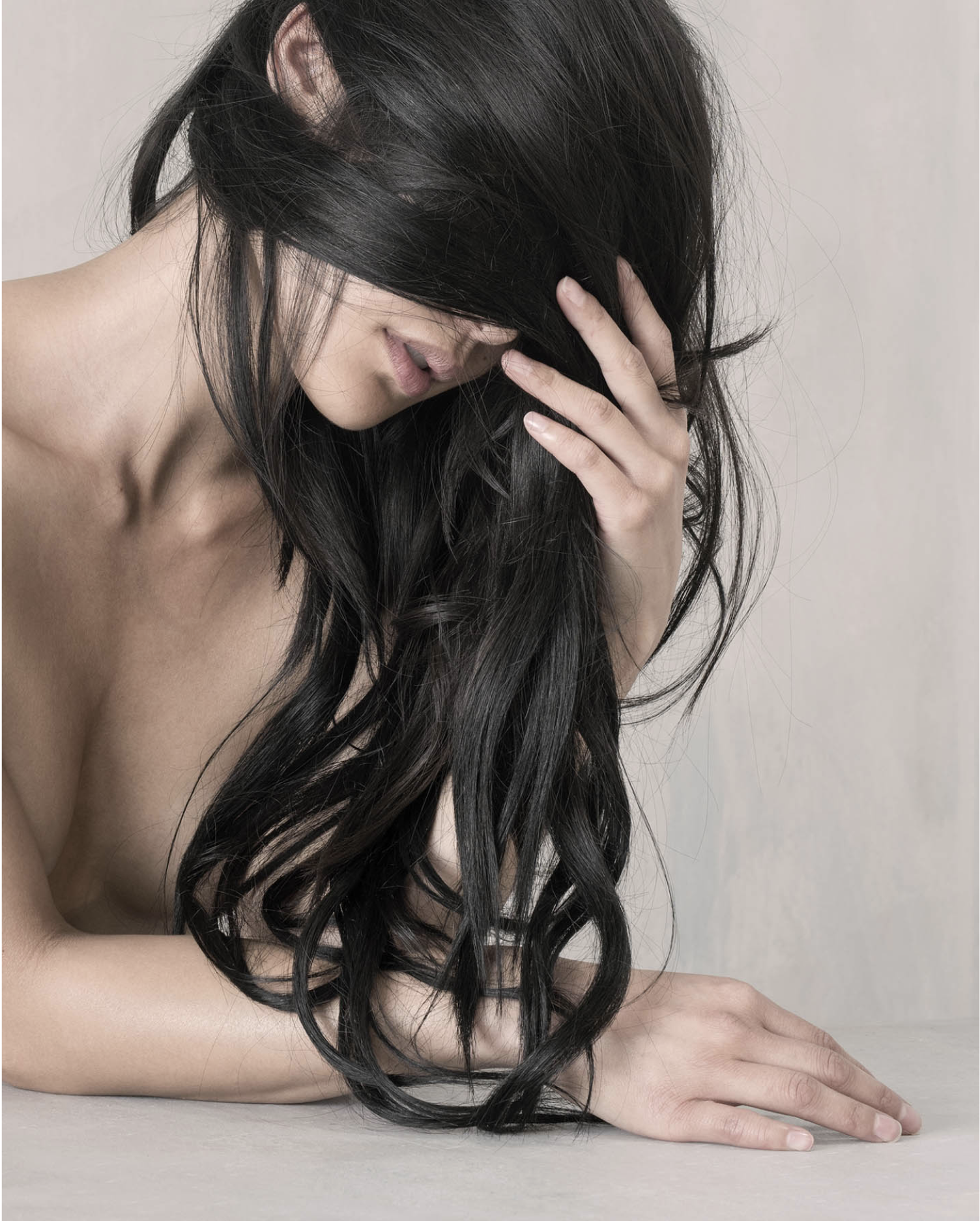 A woman with long dark hair covering part of her face, resting her head on one hand and lying on a flat surface, with the other hand on the surface.