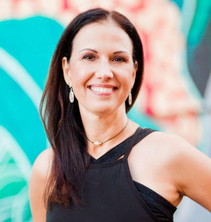 A woman with long dark hair smiling, wearing earrings and a black sleeveless top, with a colorful mural background.