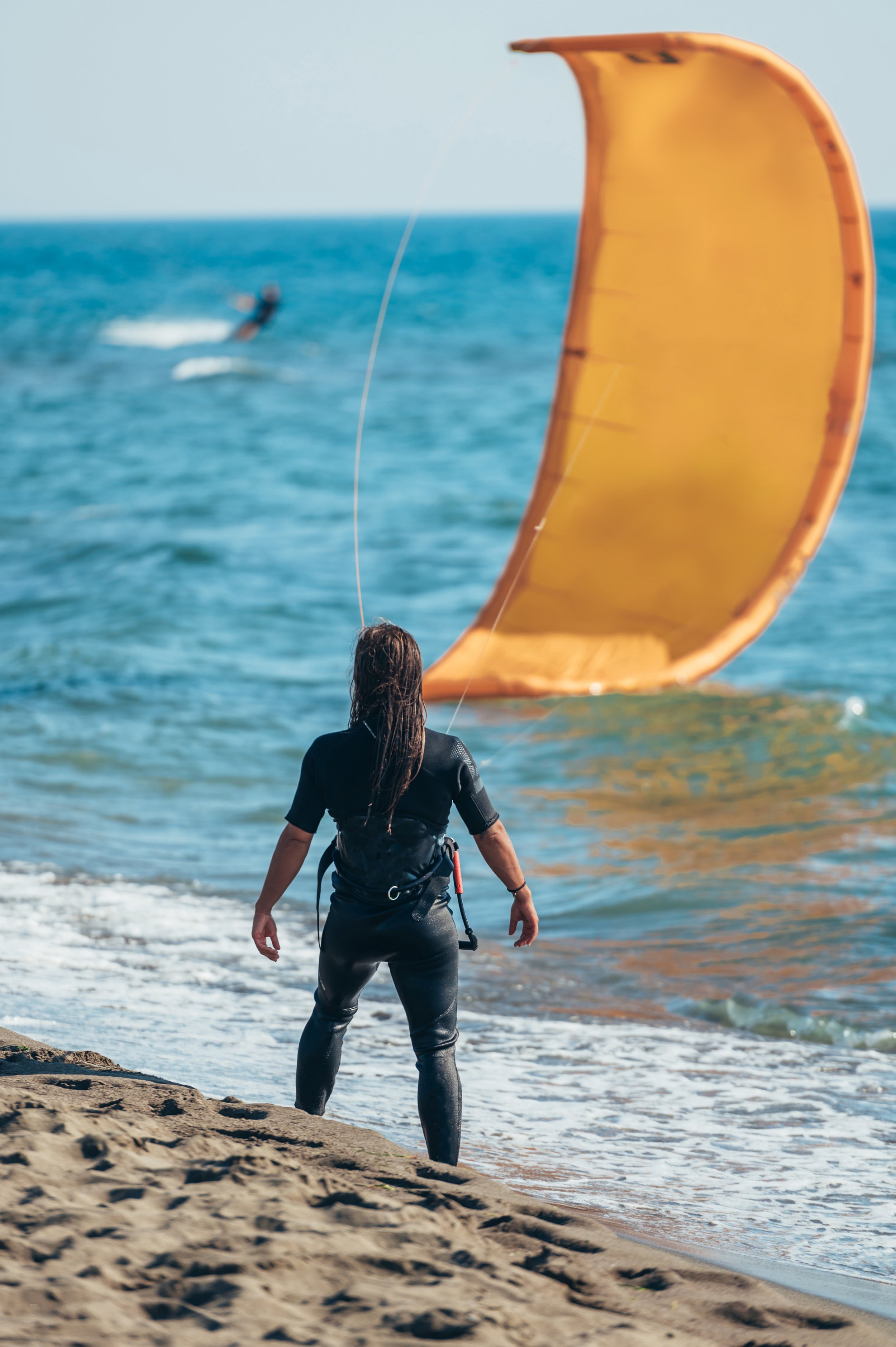 Person standing on sandy beach, preparing to launch kite surfing equipment, with a large orange kite in the water and someone kite surfing in the background.