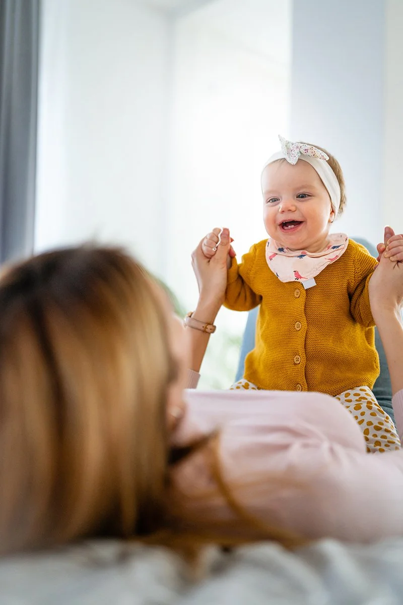 A baby girl with a white bow headband and yellow cardigan is standing and smiling while holding hands with an adult. The adult is lying on a bed with brown hair visible, and they are holding the baby's hands. The background shows a bright room with natural light.