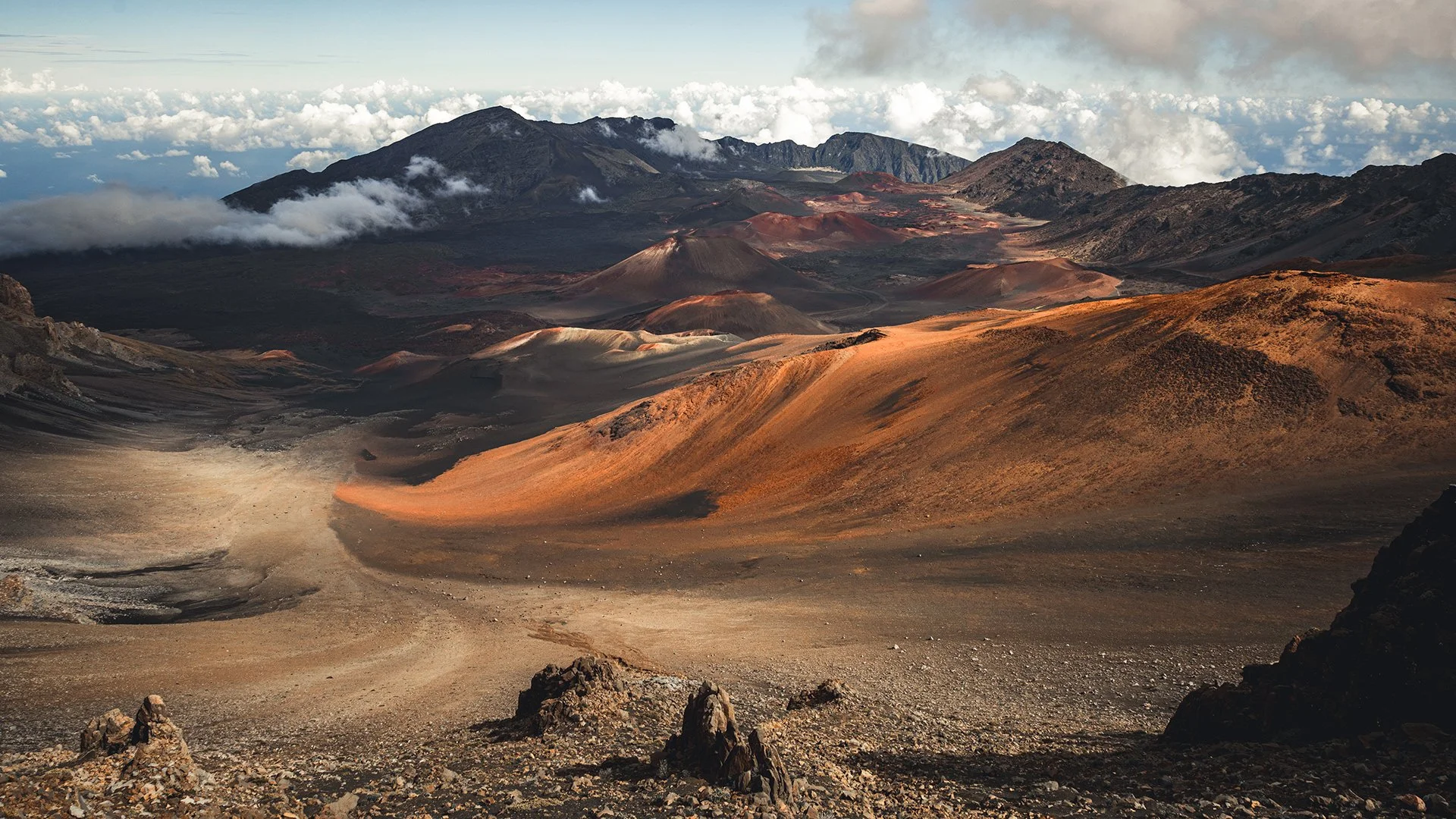 A volcanic landscape with dark, rugged mountains, colorful volcanic cones, and dry, rocky terrain under a partly cloudy sky.