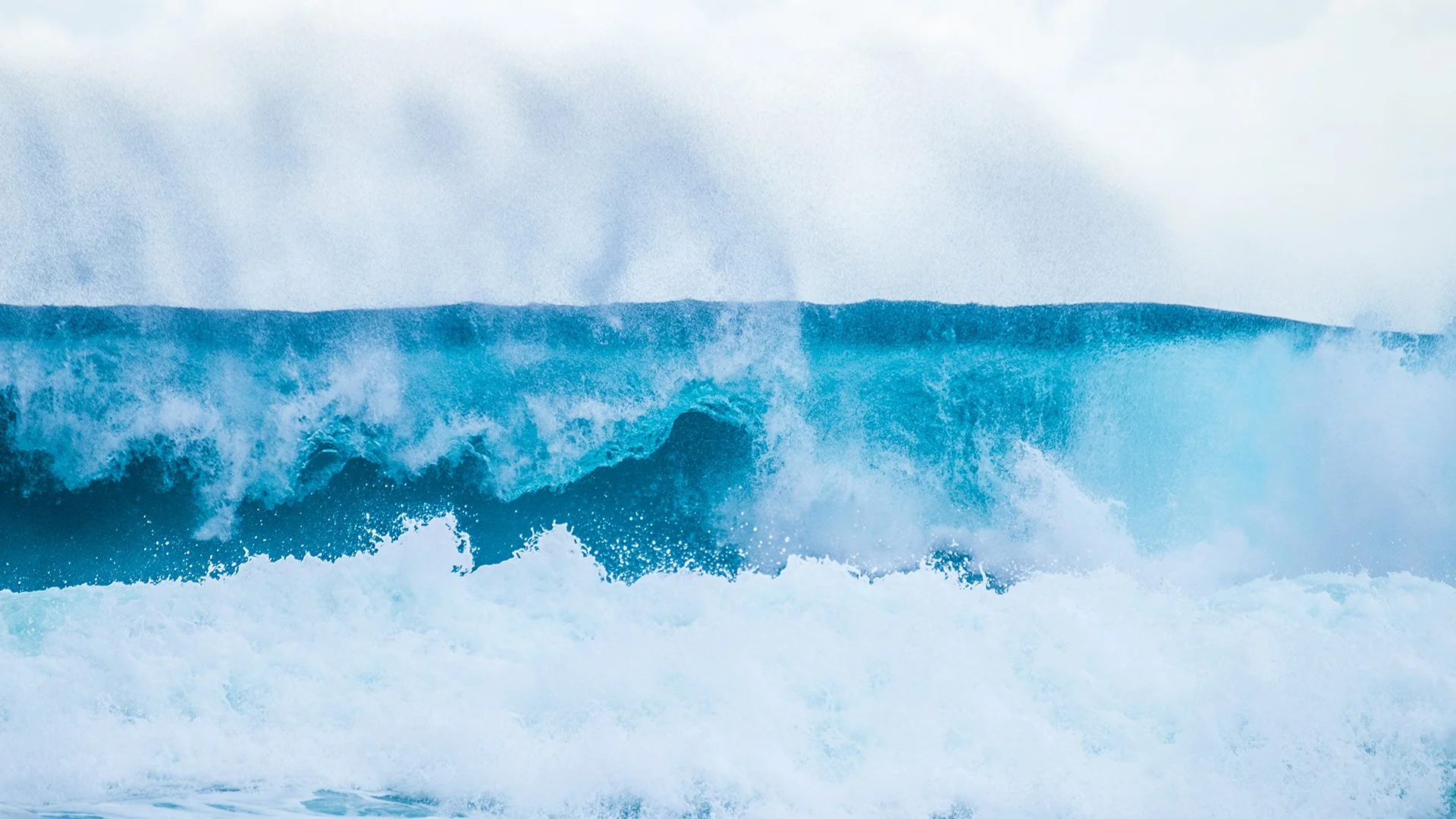 A large ocean wave with white foam and spray, under a cloudy sky.