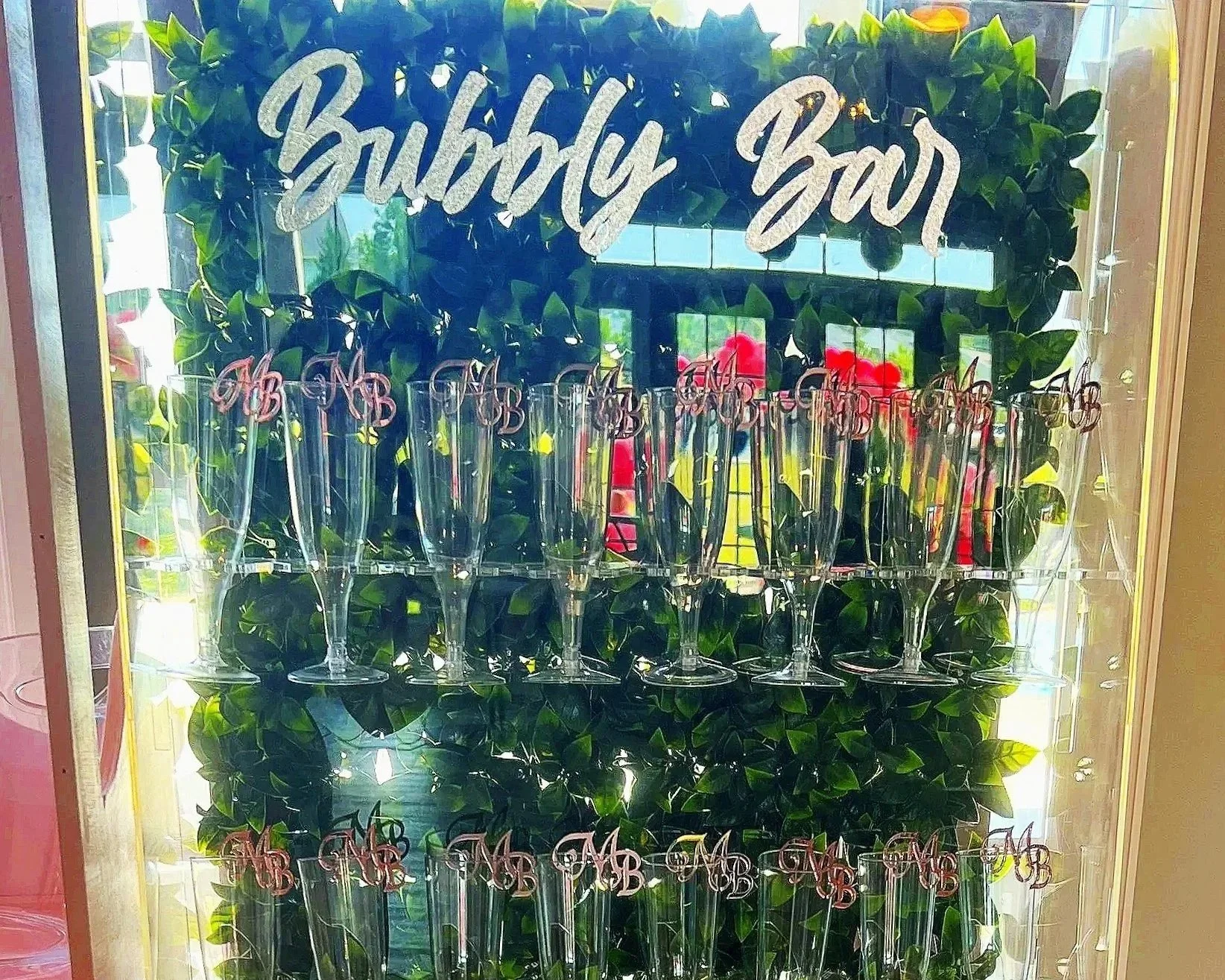 Glass display with empty champagne flutes and a decorative sign that reads 'Bubbly Bar' above it, with a background of green foliage and red flowers visible through the glass.