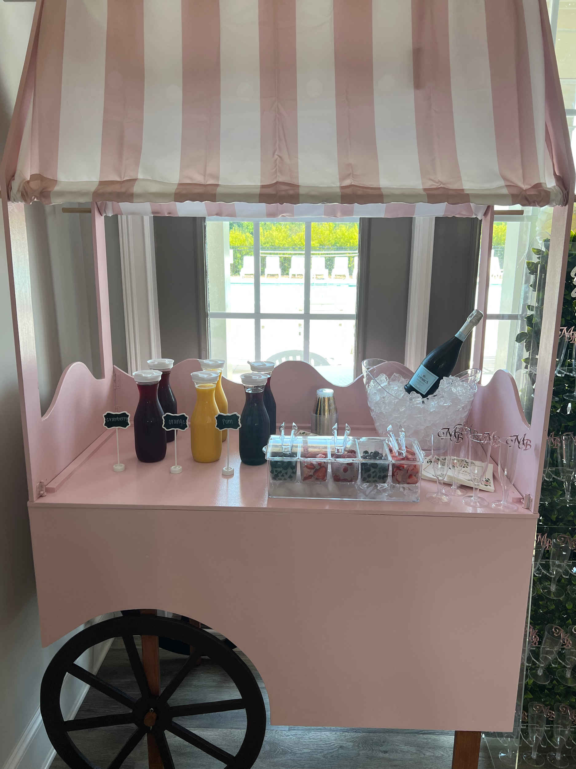 Pink mobile drink cart with striped canopy, various fruit juices, a bottle of champagne in a bucket, and cups, set by a window.