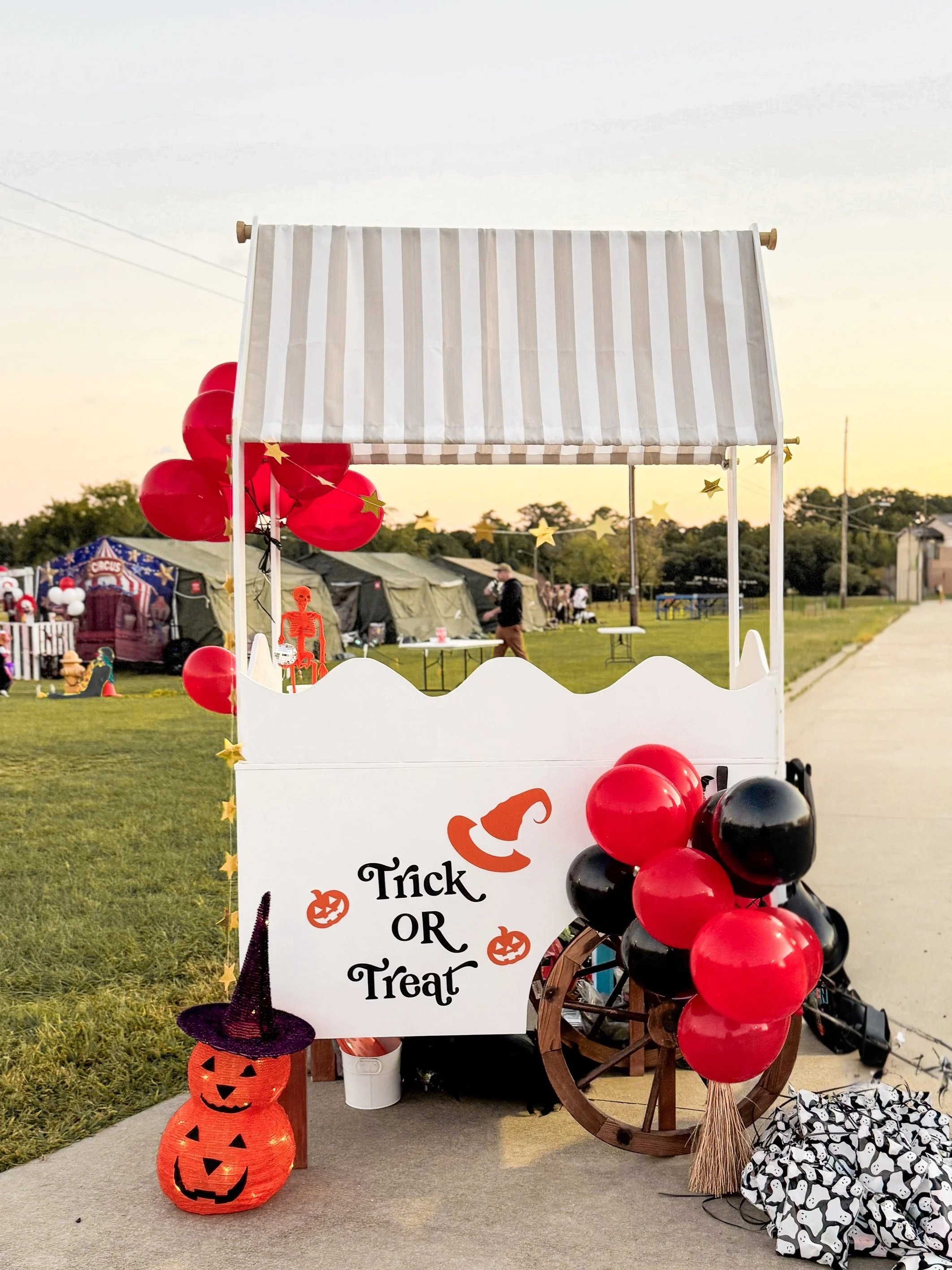 A decorated Halloween-themed small cart with a sign that reads 'Trick or Treat,' red and black balloons, a witch hat, paper pumpkins, and plastic skeletons, set outdoors at dusk.