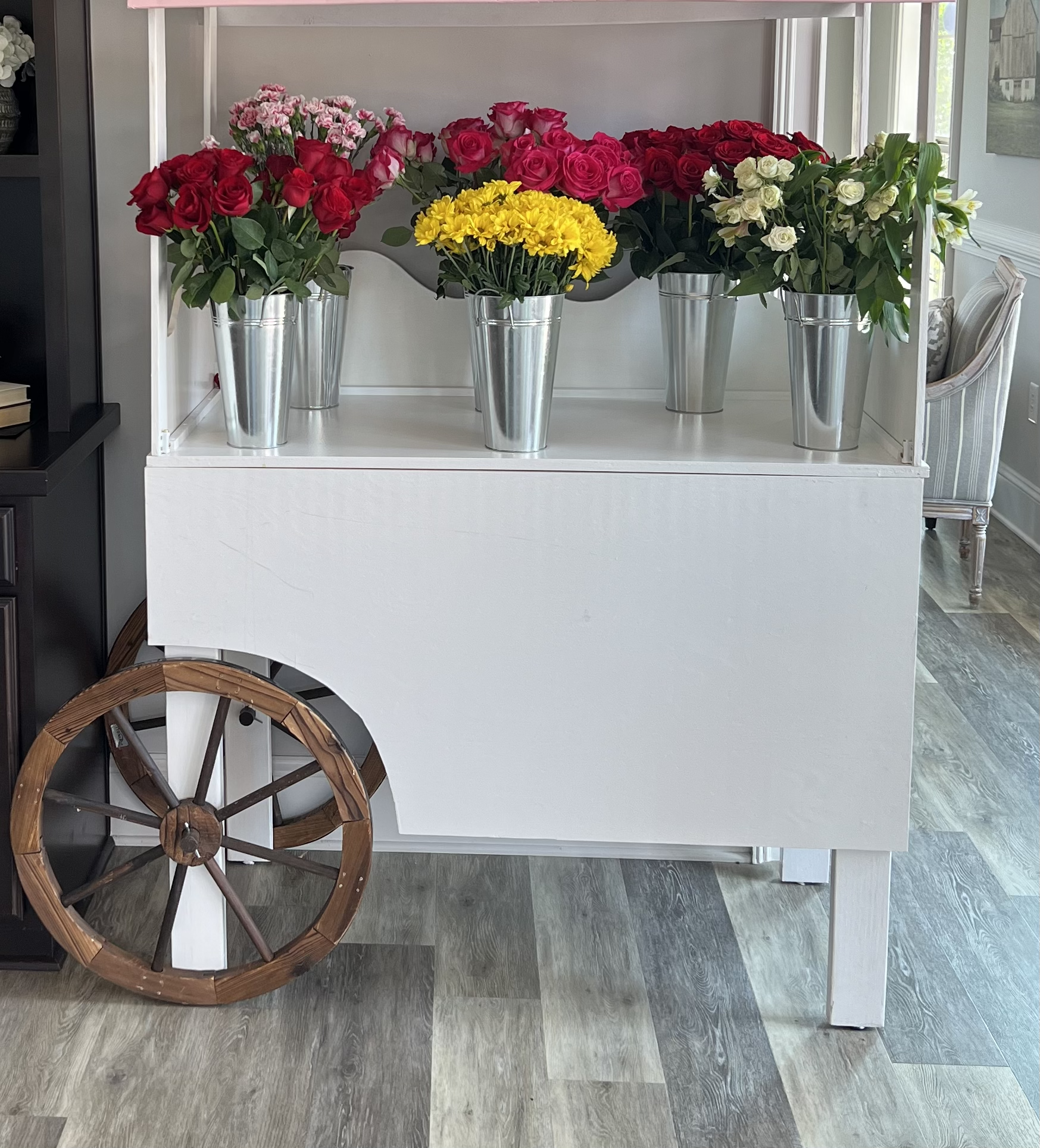 A mobile display cart with a white wood frame and a large wheel on the left side, holding five silver metal buckets filled with pink, red, yellow, and white flowers, situated on a wood floor in a room with a chair and framed artwork visible in the ba