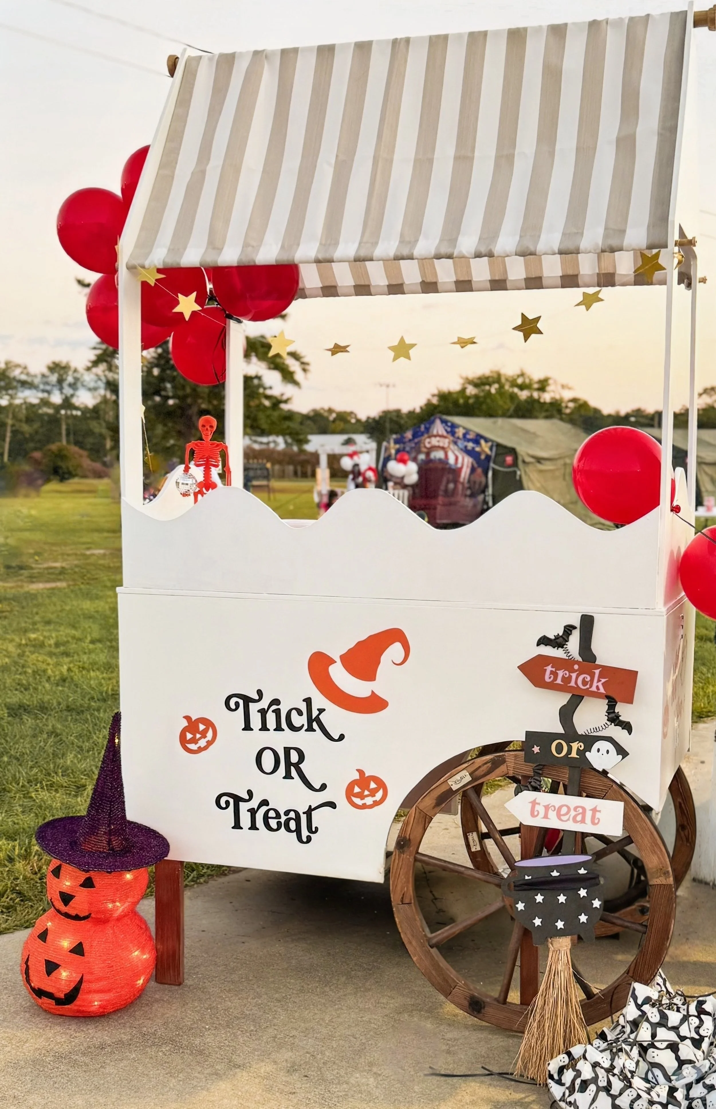 A Halloween decorated cart with red balloons, a small skeleton figurine, pumpkins with a witch hat, and Halloween signs, set outdoors at sunset.