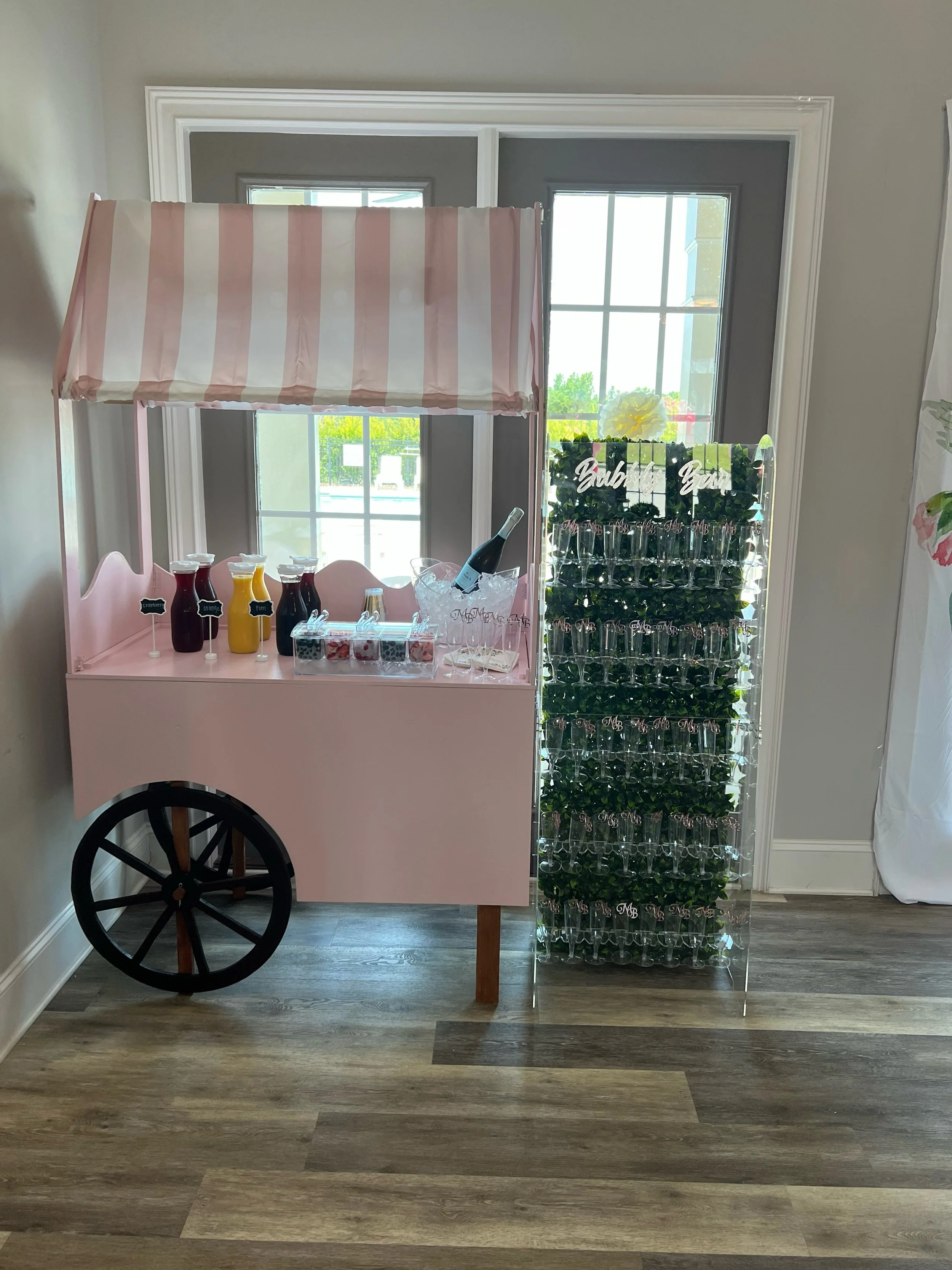Pink carnival-style cart with a striped canopy, holding juice bottles, a champagne bottle, and small cups, alongside a clear stand with hanging glass tubes and artificial greenery, on a wooden floor near windows.