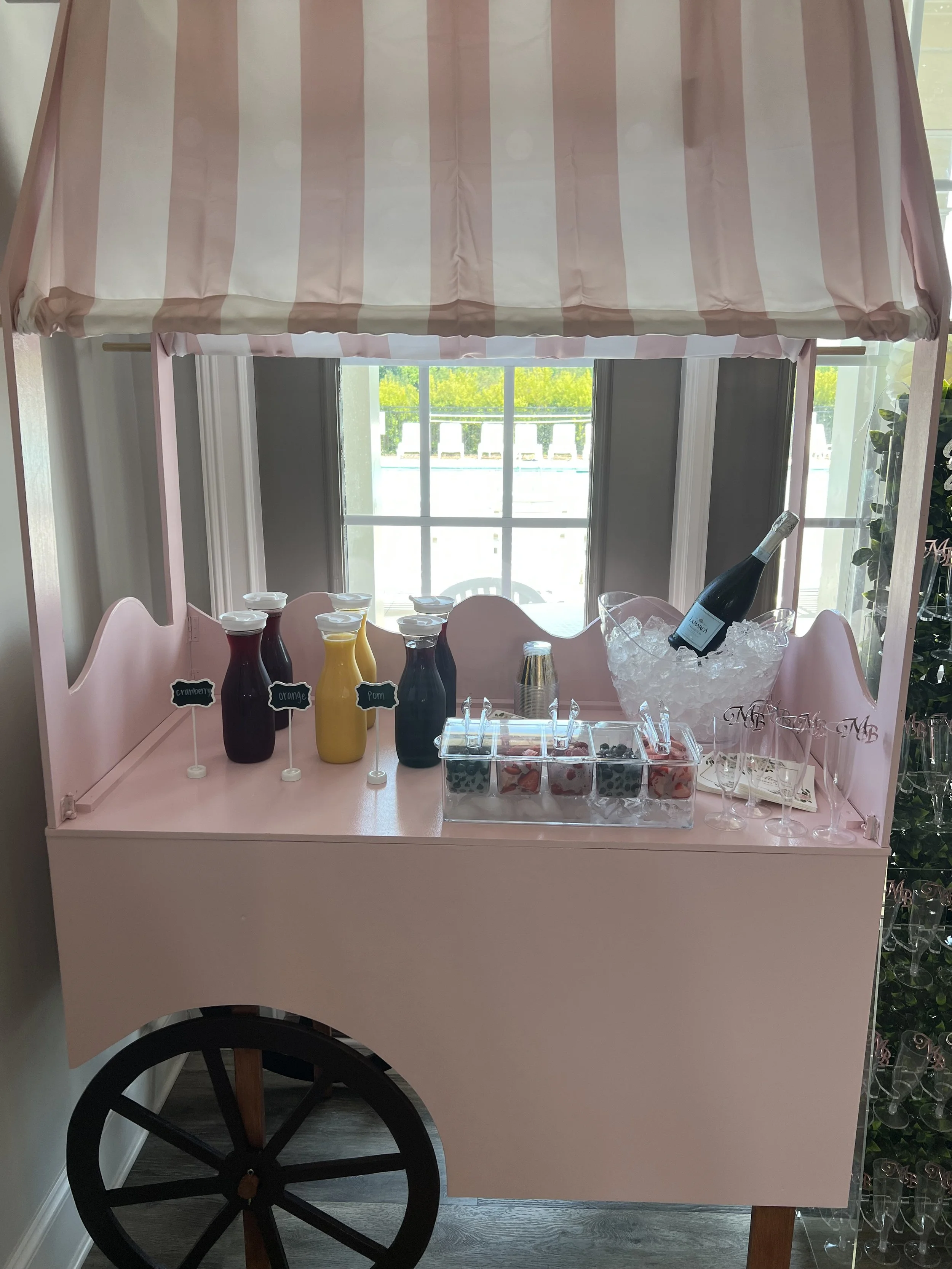 Pink cart with a striped canopy serving beverages, including juice bottles labeled lemon, orange, and pom, and a champagne bottle in ice, with small cups and covered containers of garnishes, set near a window in a bright room.