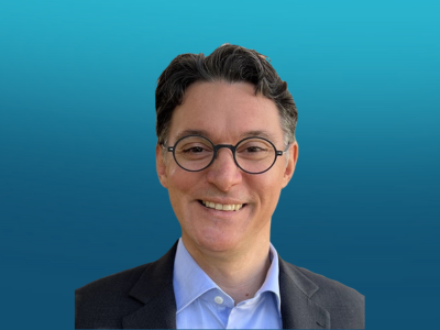 Portrait of a smiling man with glasses, dark hair, wearing a suit and light blue shirt, against a blue background.