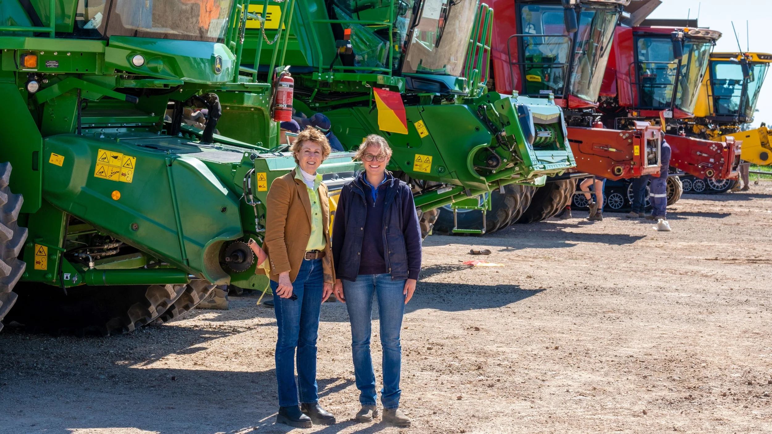 Two women standing in front of a row of large green, red, and yellow construction or farming equipment outdoors.