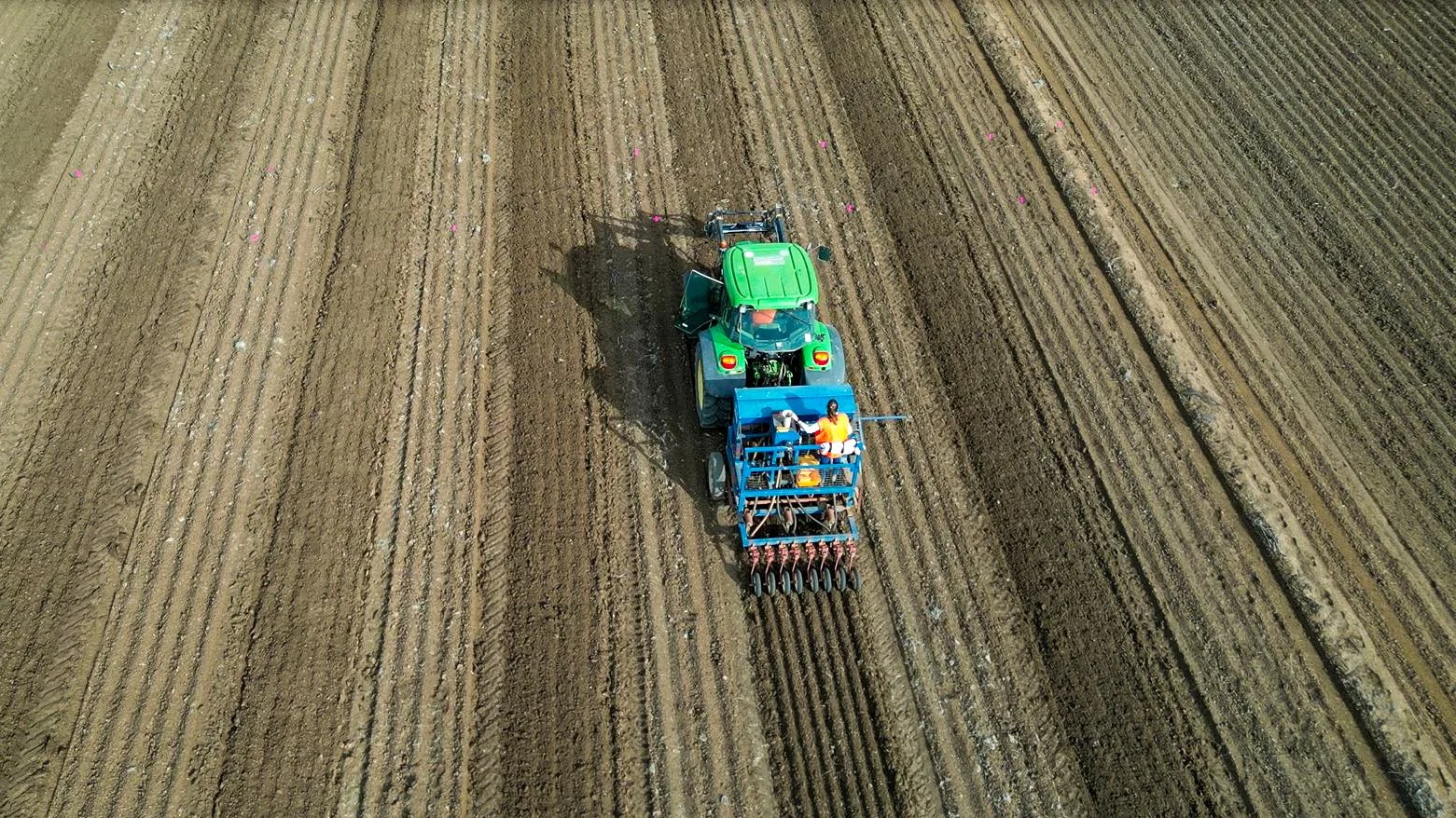 An aerial view of a green tractor working on tilled soil in a farm field, with a blue planting attachment and several workers on it.
