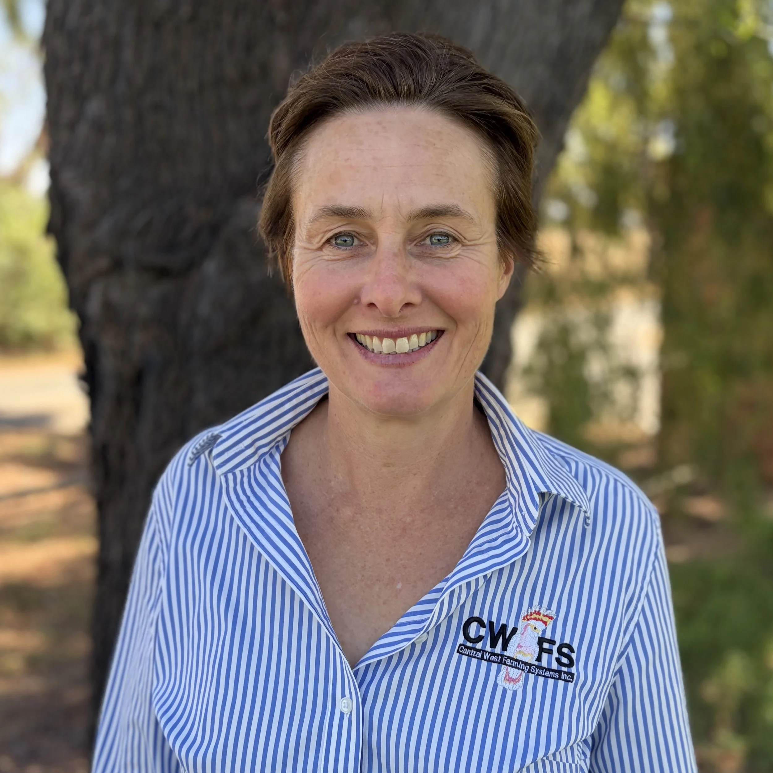 A smiling woman with short brown hair wearing a blue and white striped shirt with a logo that says 'Central West Fanning Systems Inc.' standing outdoors in front of a tree with blurred green and yellow foliage.