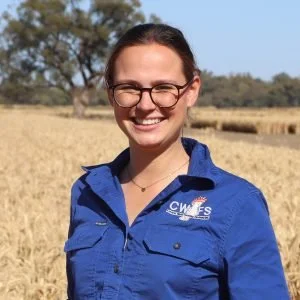 A woman smiling outdoors in a field, wearing glasses and a blue jacket with a logo.