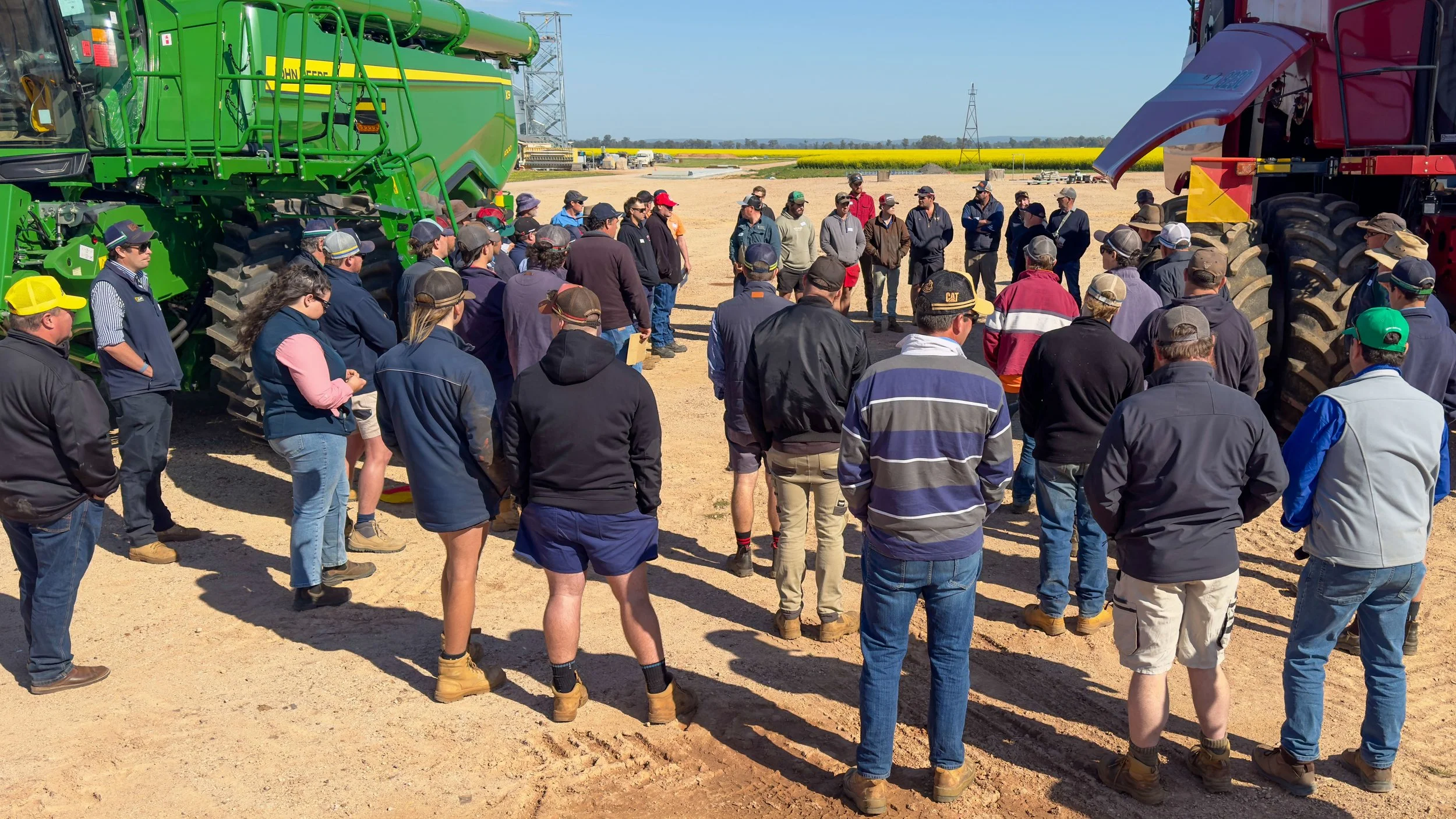 A group of people gathered outdoors in an open dirt area, surrounded by large green and red agricultural machinery, possibly at a farm or equipment demonstration event.