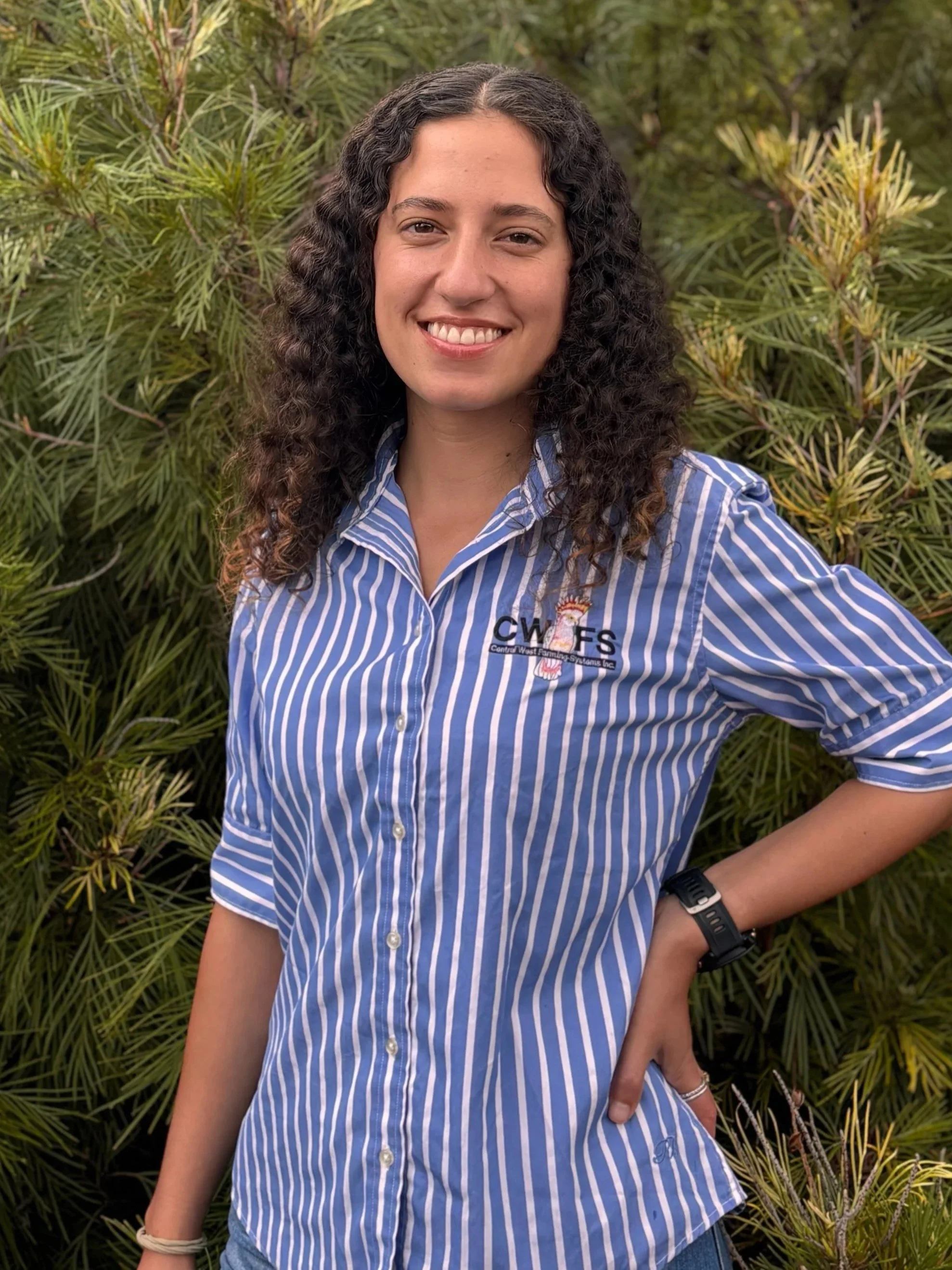 A young woman with curly dark hair, wearing a blue and white striped shirt, standing outdoors in front of green foliage, smiling at the camera.
