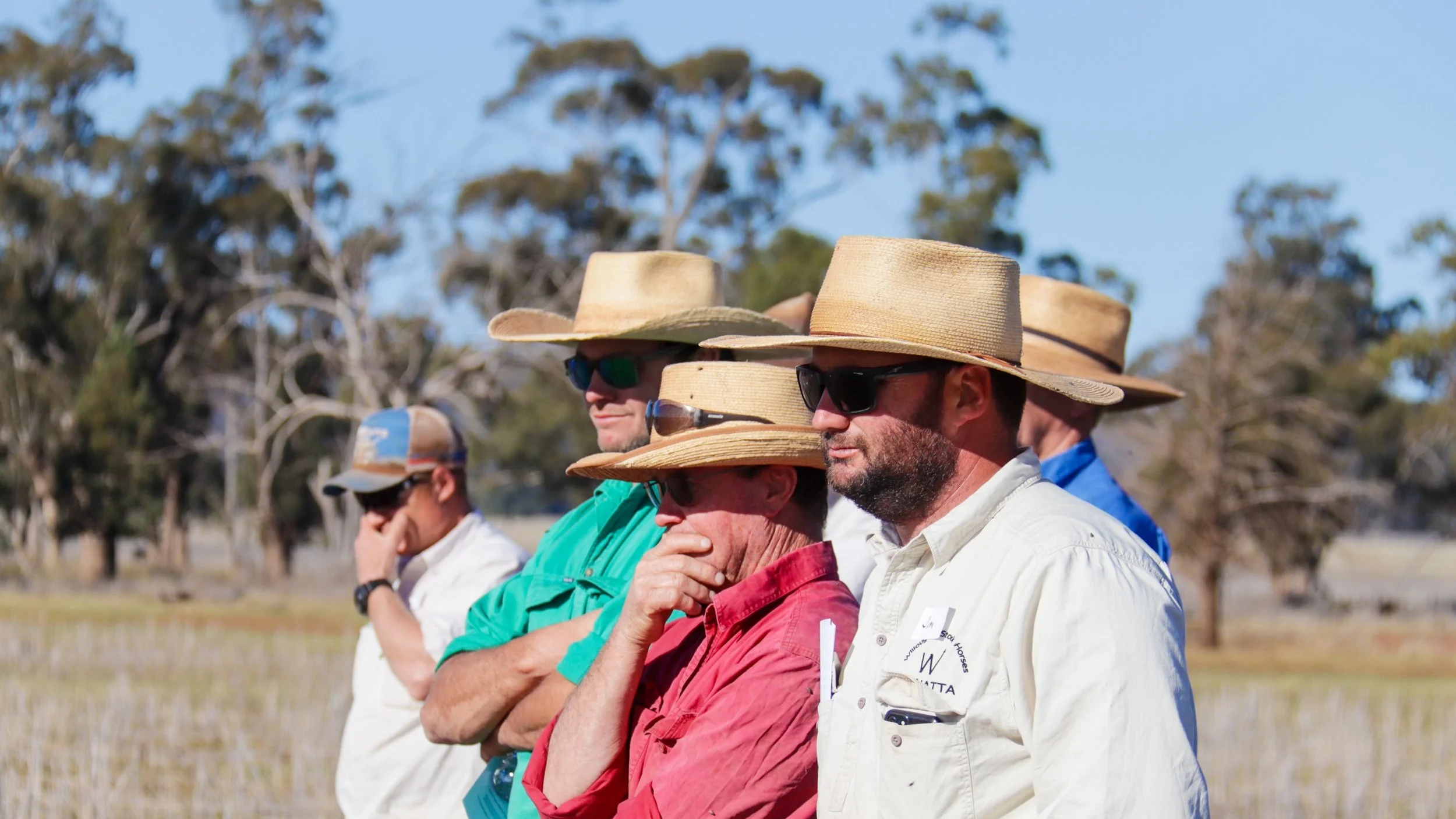 Group of men wearing wide-brimmed hats and sunglasses, standing outdoors in a field, with trees and blue sky in the background.