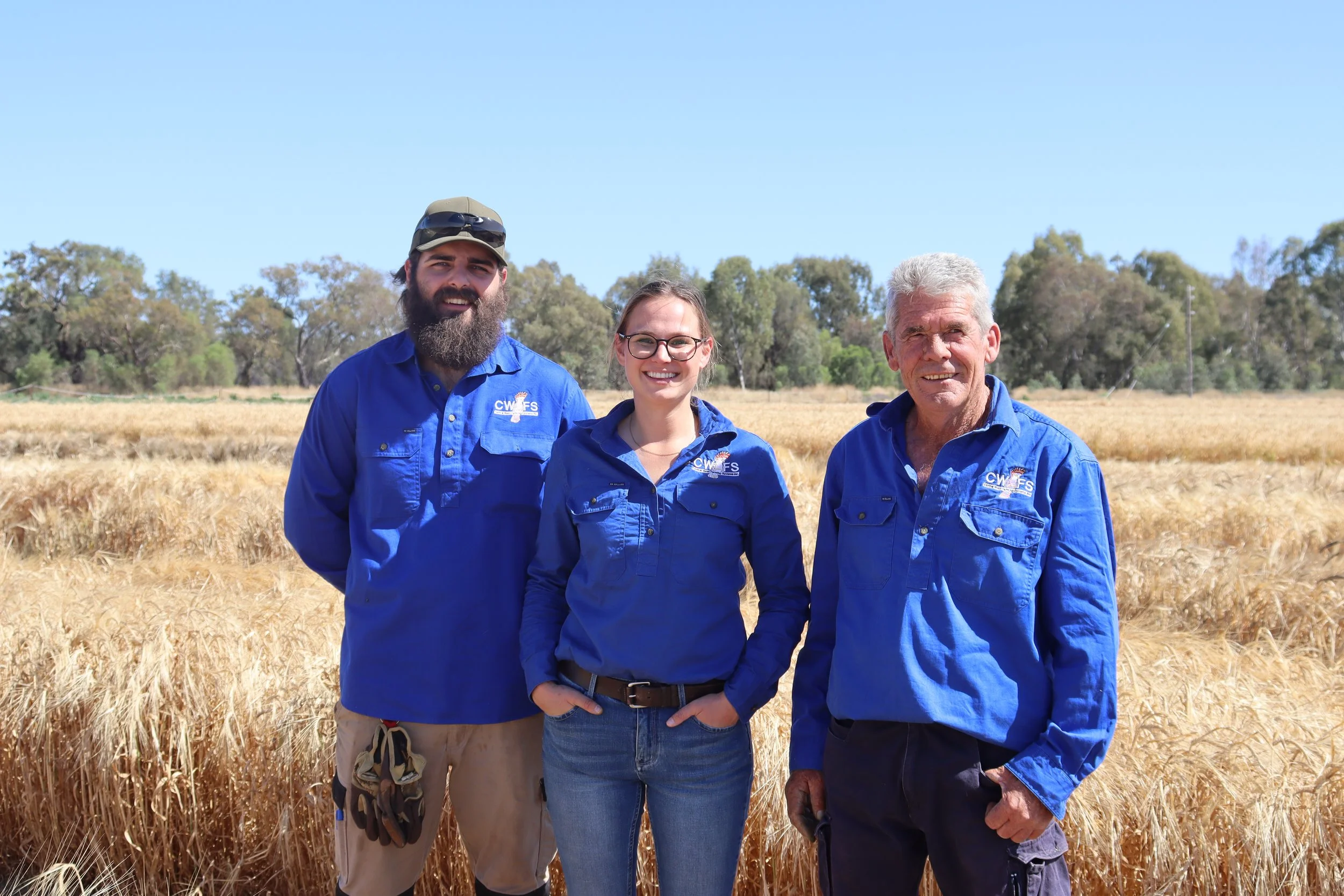 Three people standing in a wheat field, smiling at the camera, wearing blue shirts with a logo, and with trees in the background.