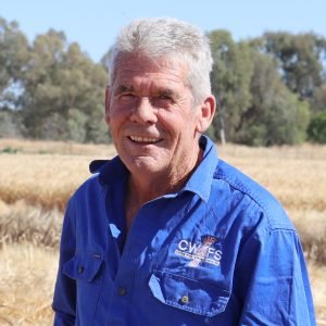 A smiling man with white hair wearing a blue shirt standing outdoors in a field with trees in the background.