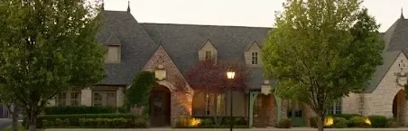 A large house with a stone exterior, multiple gabled roofs, and an arched entrance, surrounded by green trees and shrubs.