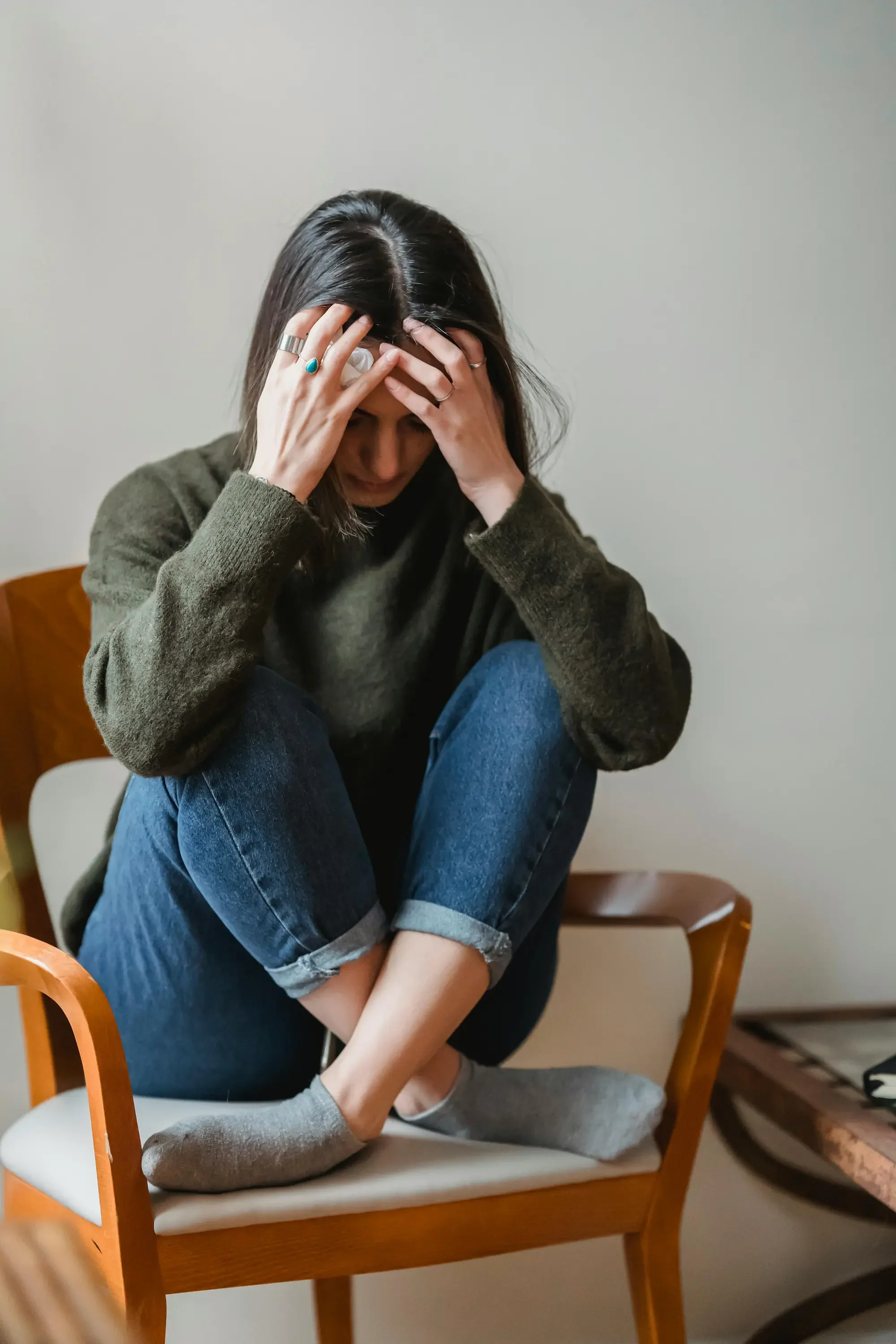 A woman sitting in a chair looking down with her hands on her head, illustrating feelings of overwhelm or anxiety in a therapy setting.