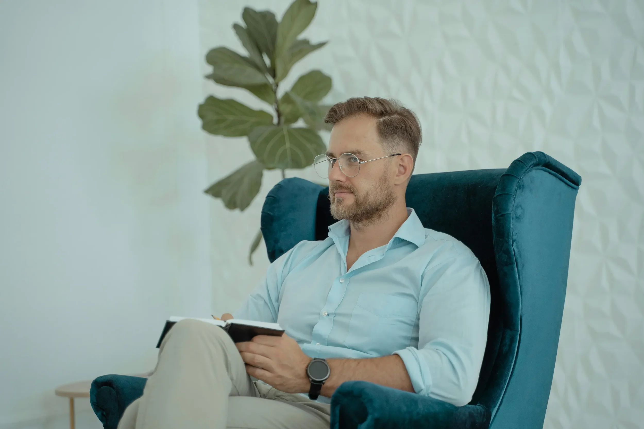 A male therapist with glasses and a beard sitting in a comfortable teal wingback chair, holding a notebook and listening attentively.