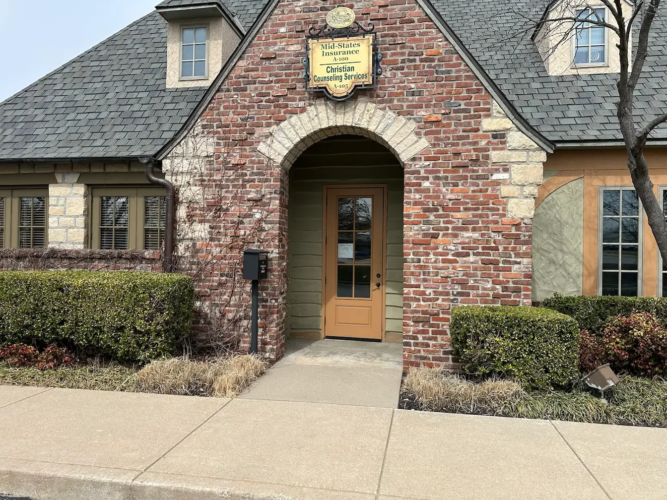 A welcoming brick building entrance with an arched doorway, manicured green bushes, and a clear sidewalk leading to the counseling office.